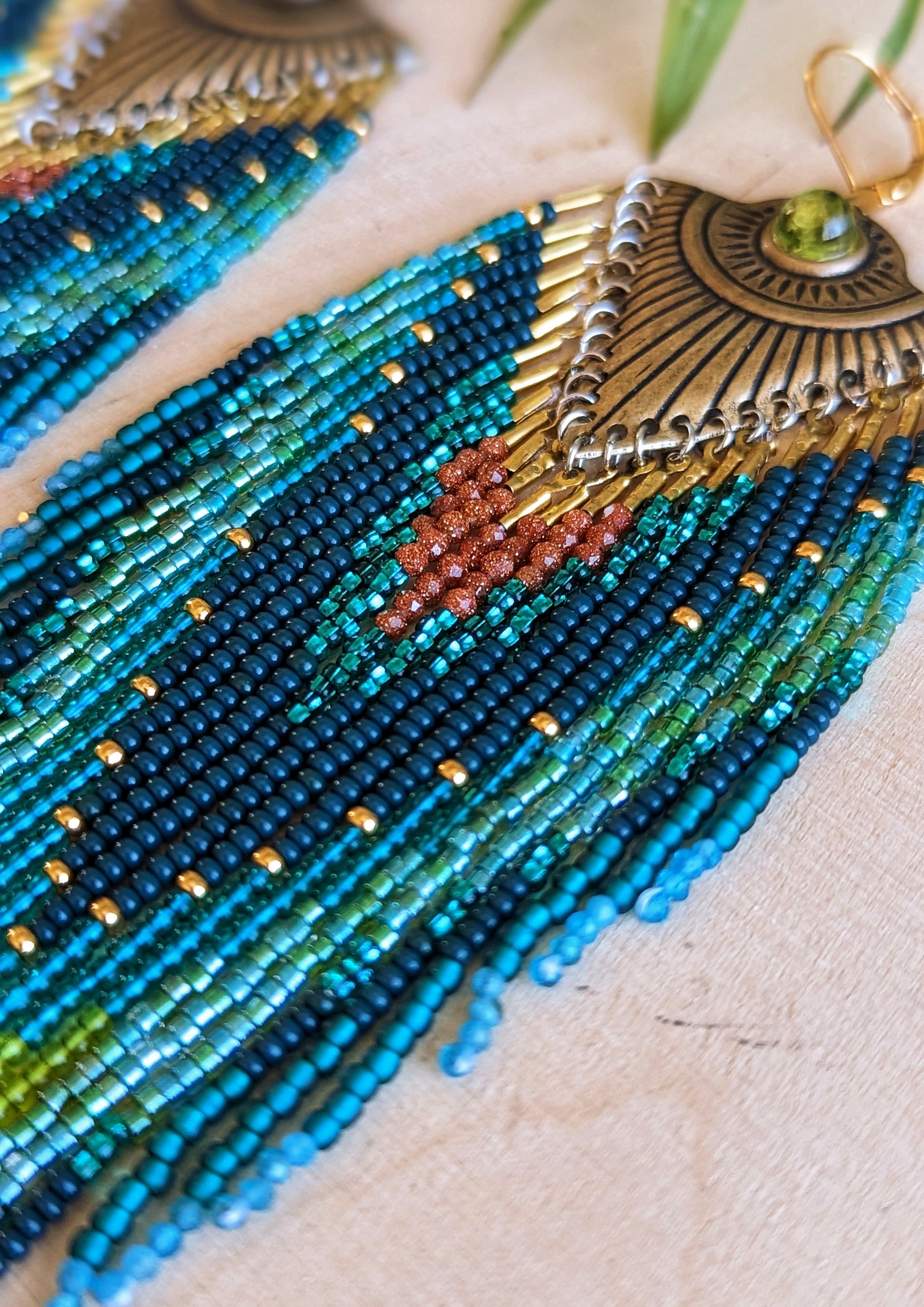 Pair of long beaded earrings featuring intricate patterns in deep blue, lime green, teal, and rust, with a brass sunburst accent at the top, and a dark green stone embedded, lying on a wooden surface.