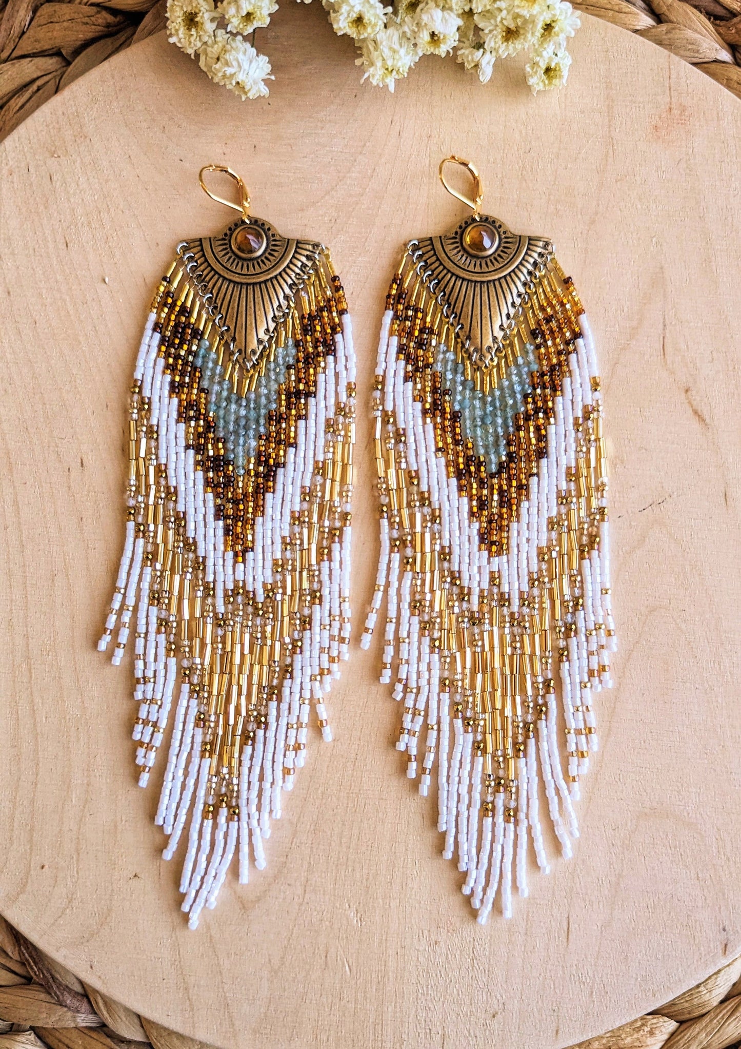 Moon & Milk’s Tonatiuh earrings displayed on wood, featuring white, amber, gold, and apatite stone beadwork with a radiant brass sunburst medallion at the top.