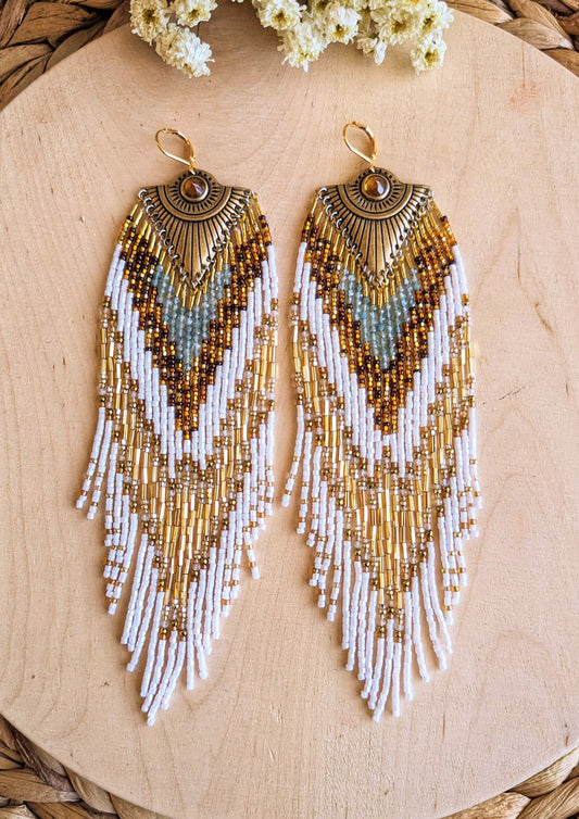 Moon & Milk’s Tonatiuh earrings displayed on wood, featuring white, amber, gold, and apatite stone beadwork with a radiant brass sunburst medallion at the top.
