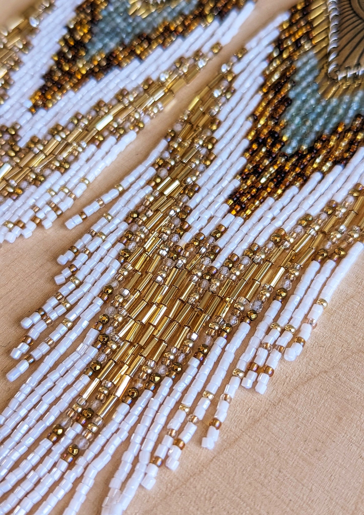 Moon & Milk’s Tonatiuh earrings displayed on wood, featuring white, amber, gold, and turquoise beadwork with a radiant brass sunburst medallion at the top.