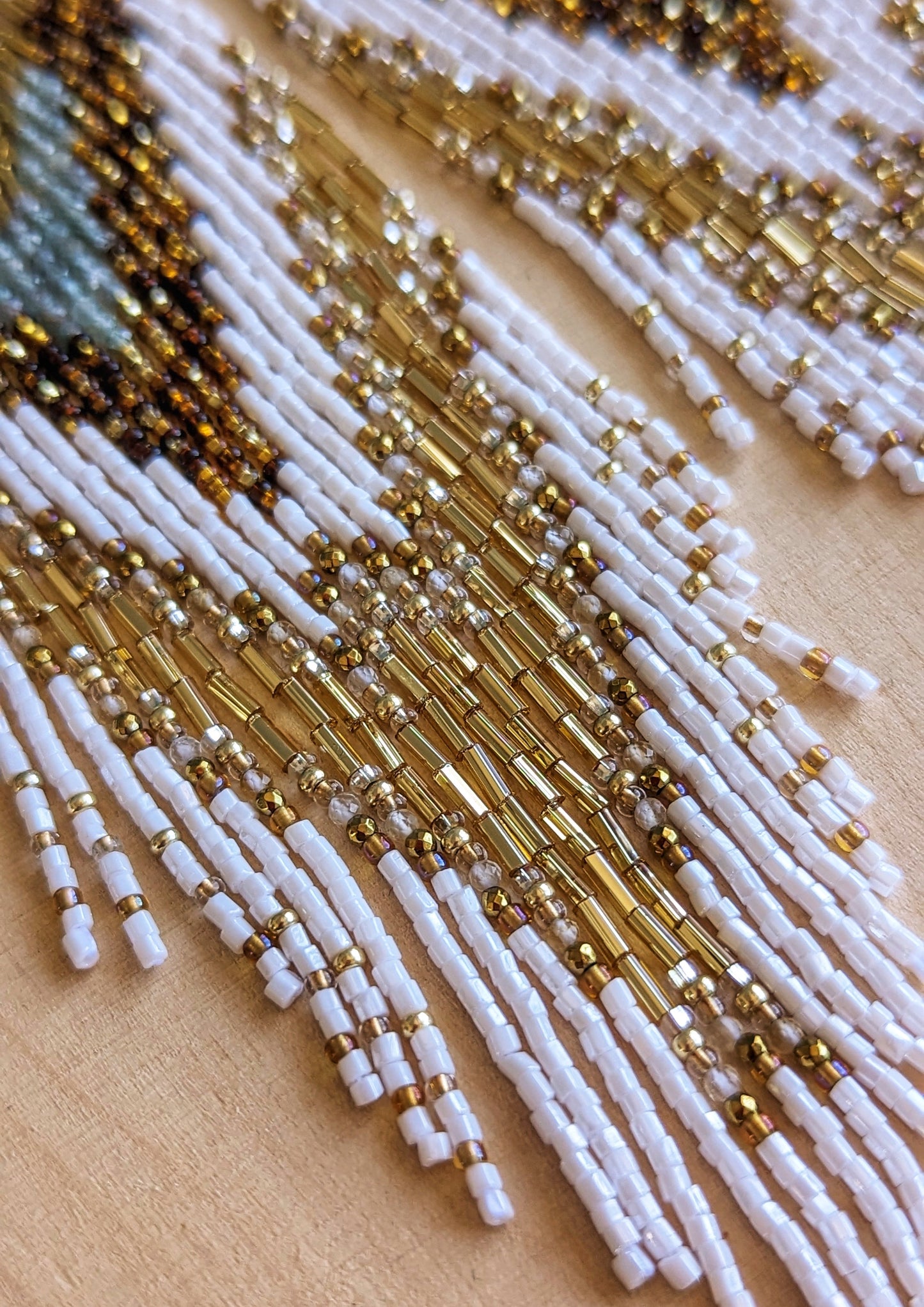 Close-up of beaded earrings displayed on wood, featuring white, amber, gold, and apatite stone beadwork with a radiant brass sunburst medallion at the top.
