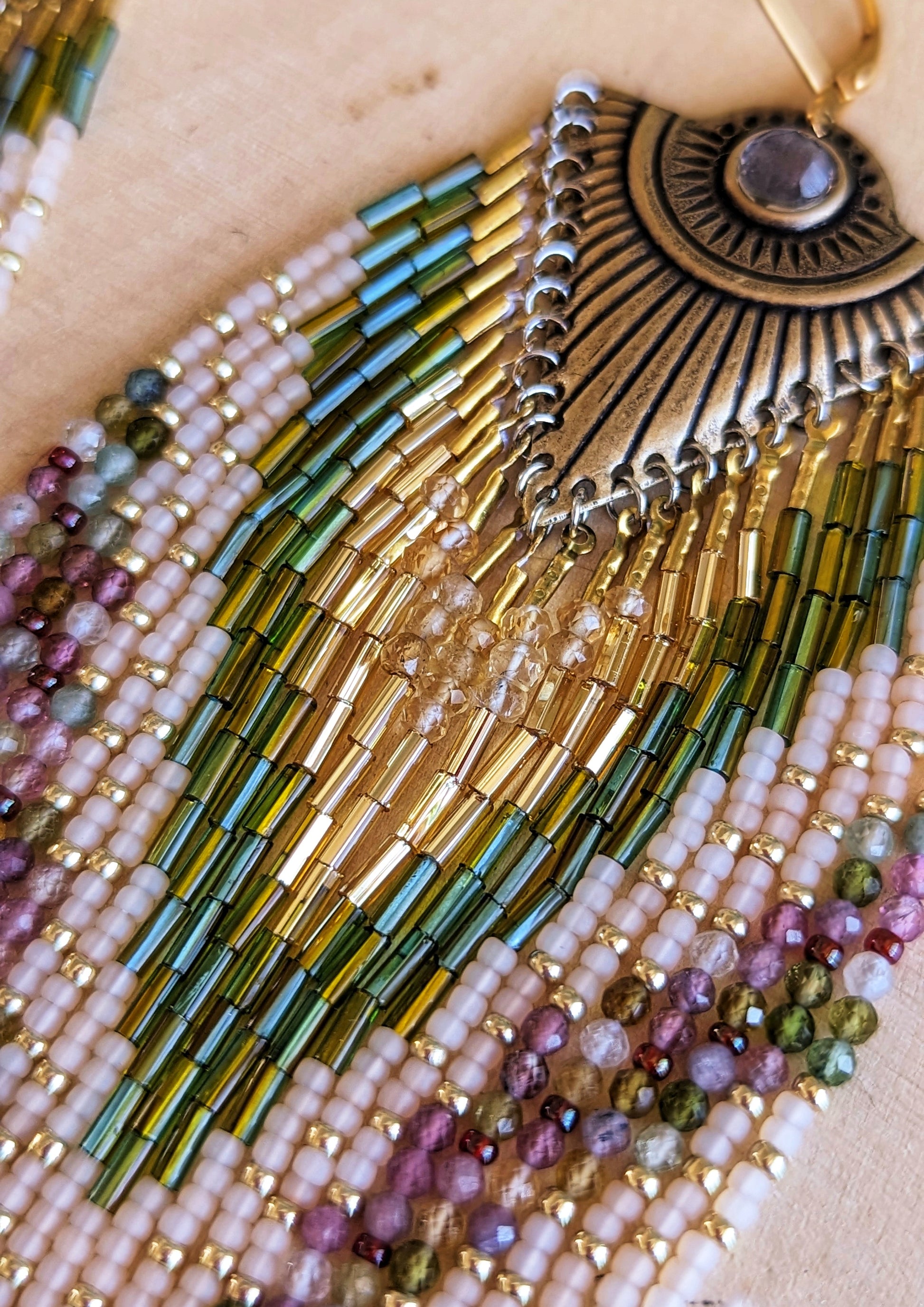 Close-up of Moon & Milk’s Xochipilli earrings showing gold, olive, blush, and citrine bead strands flowing from a sunburst brass fan with a center gem.