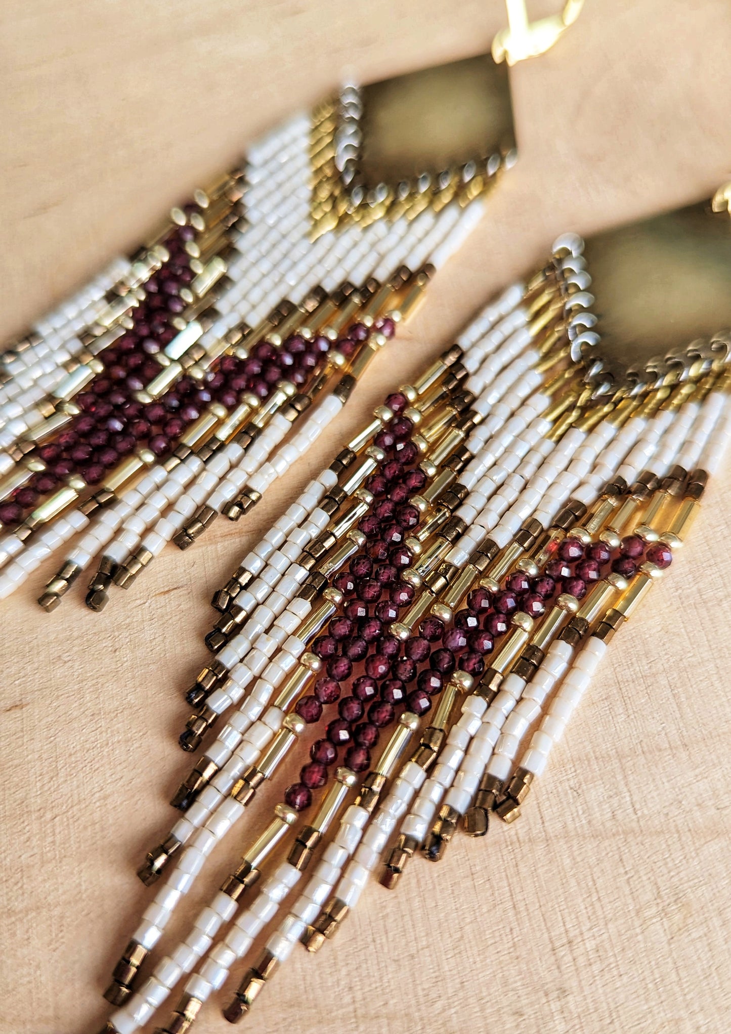 Long beaded earrings featuring garnet stones, ivory, and gold beads with brass diamond tops and chevron patterns, displayed on a wooden surface by Moon & Milk.