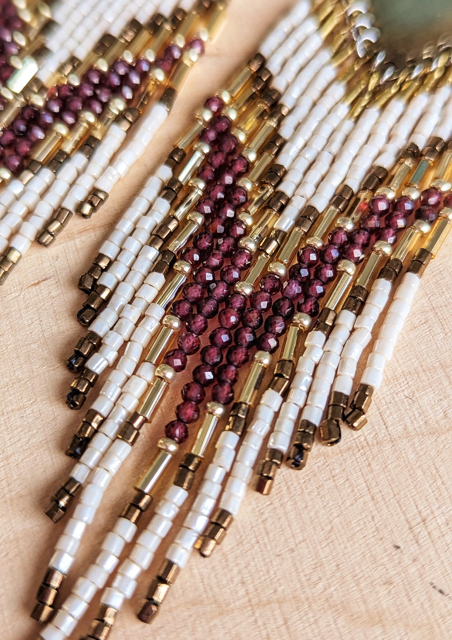 A close up of long beaded earrings featuring garnet stones, ivory, and gold beads with brass diamond tops and chevron patterns, displayed on a wooden surface by Moon & Milk.
