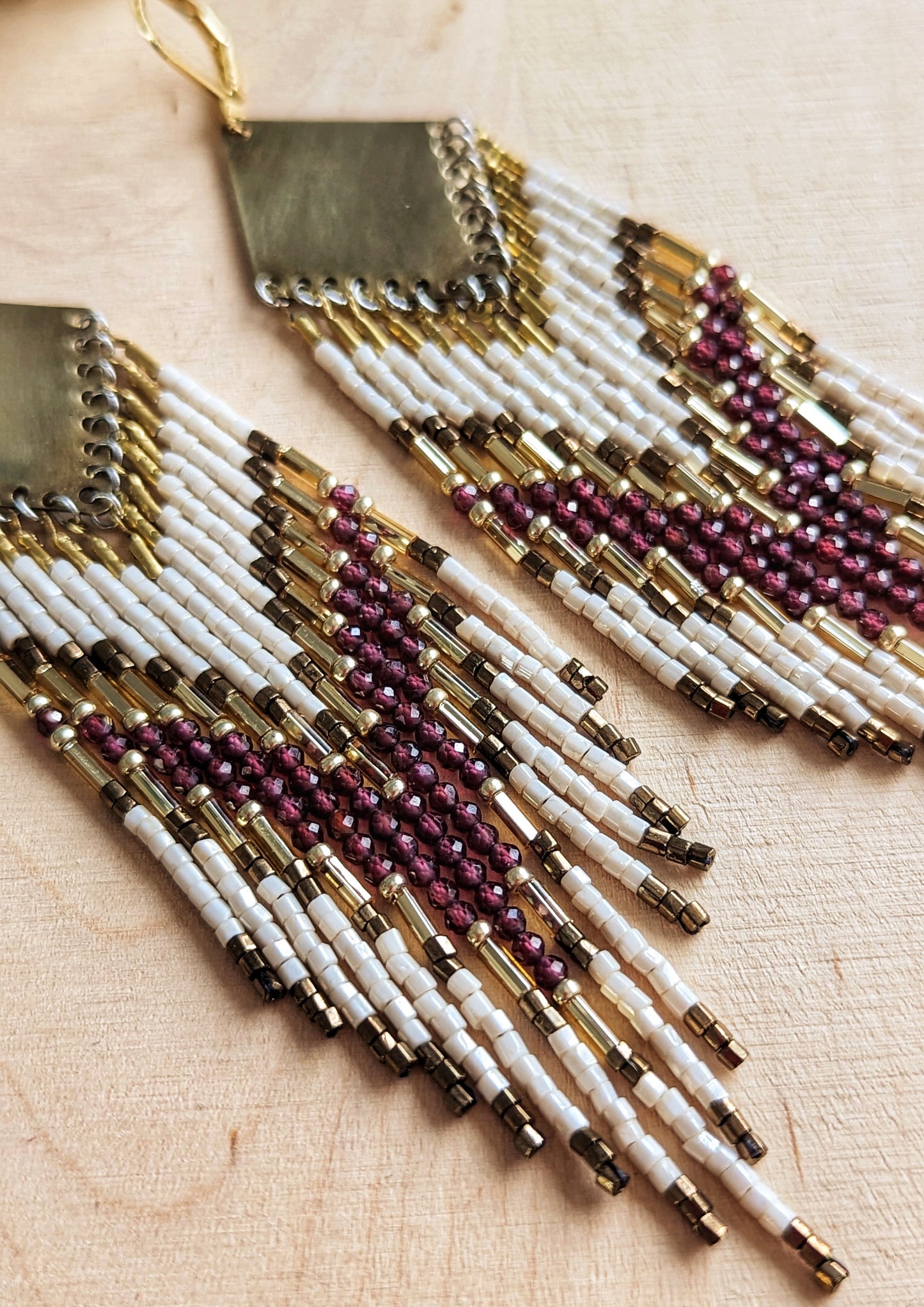 Long beaded earrings featuring garnet stones, ivory, and gold beads with brass diamond tops and chevron patterns, displayed on a wooden surface by Moon & Milk.