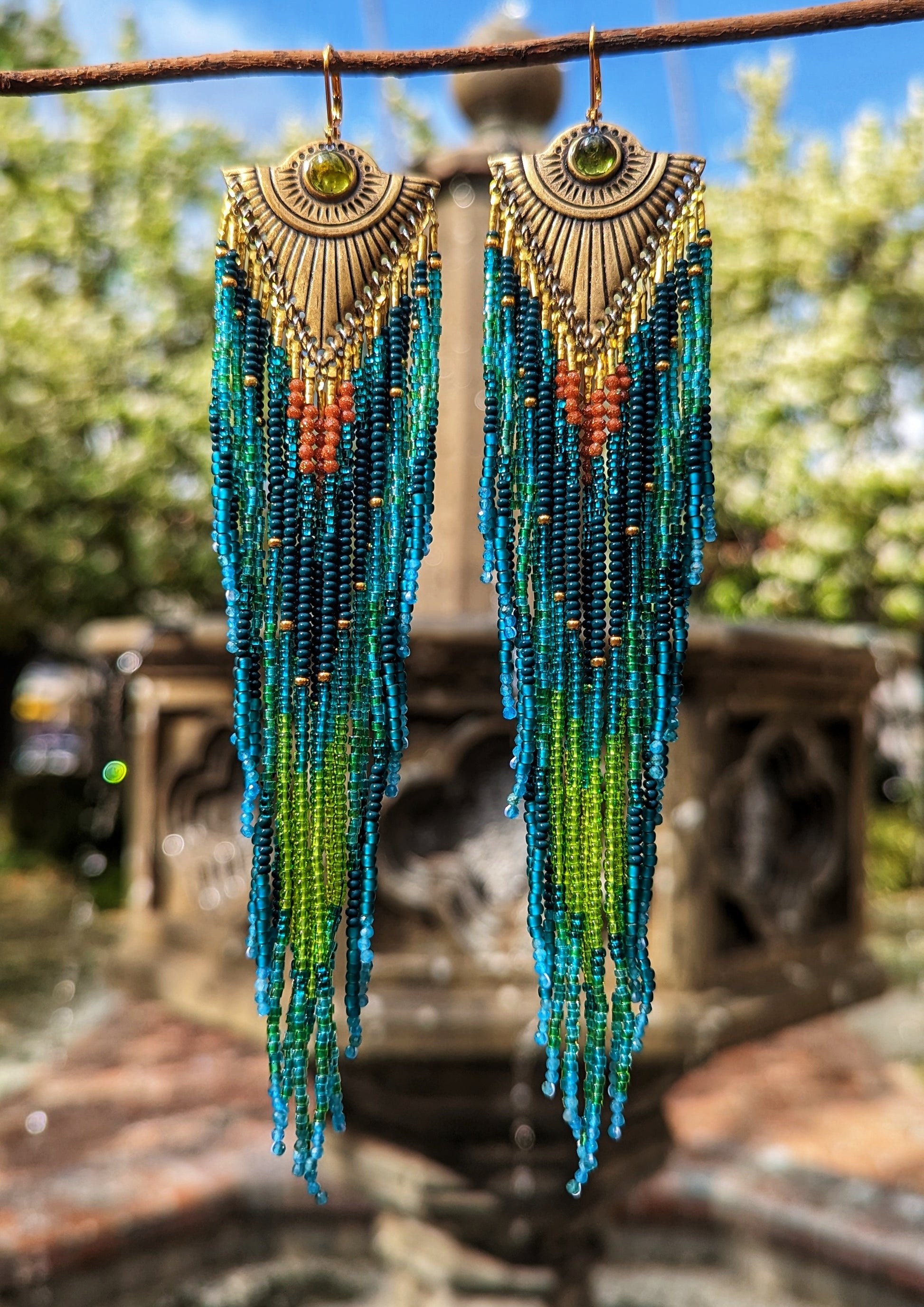 Long beaded earrings in shades of blue, green, and rust, with brass sunburst tops and embedded green stones, hanging from a branch against a blurred outdoor background.