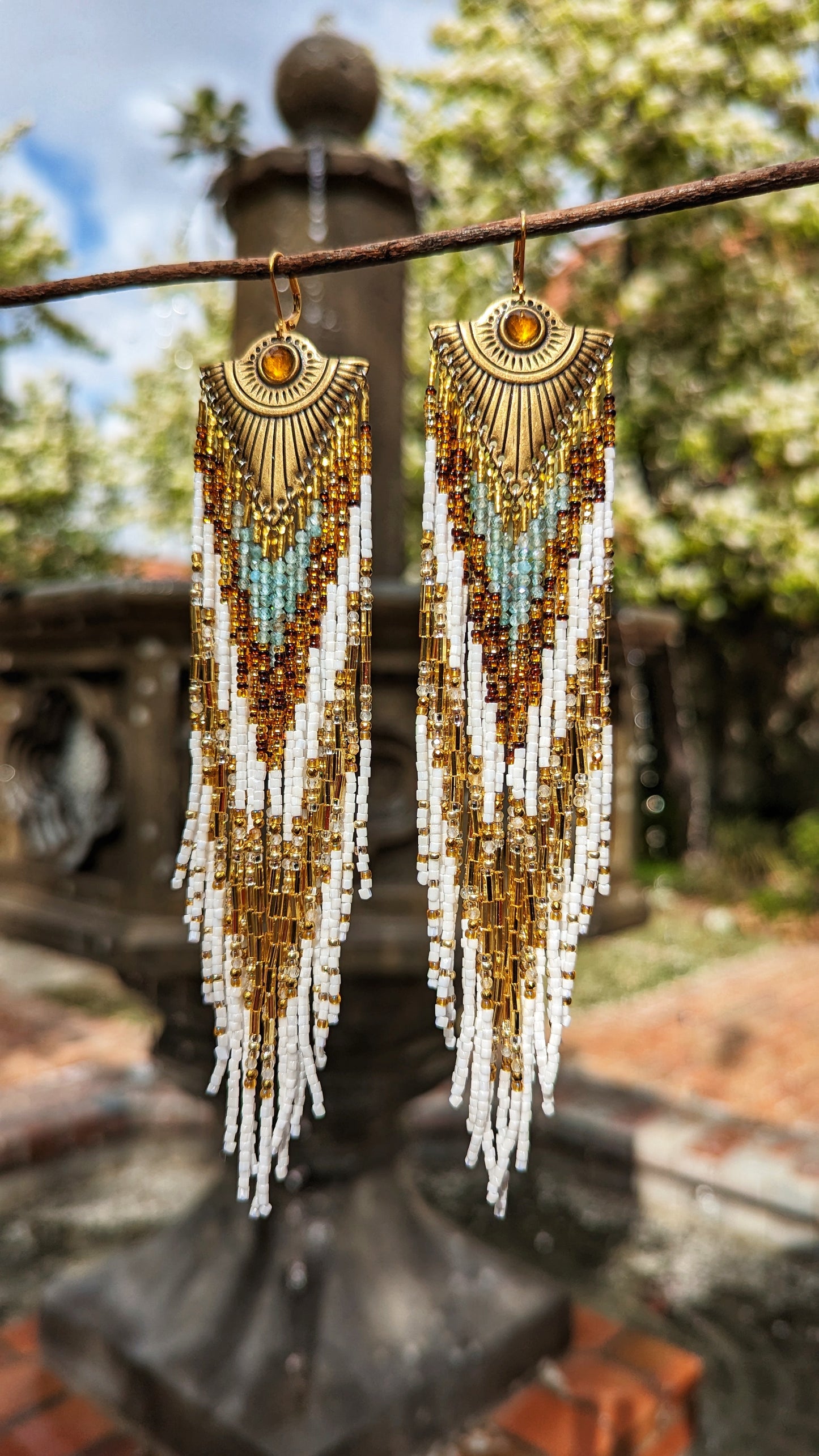 Moon & Milk’s Tonatiuh earrings hang against a garden fountain, with cascading white, gold, turquoise, and amber beads radiating from a brass sunburst base.