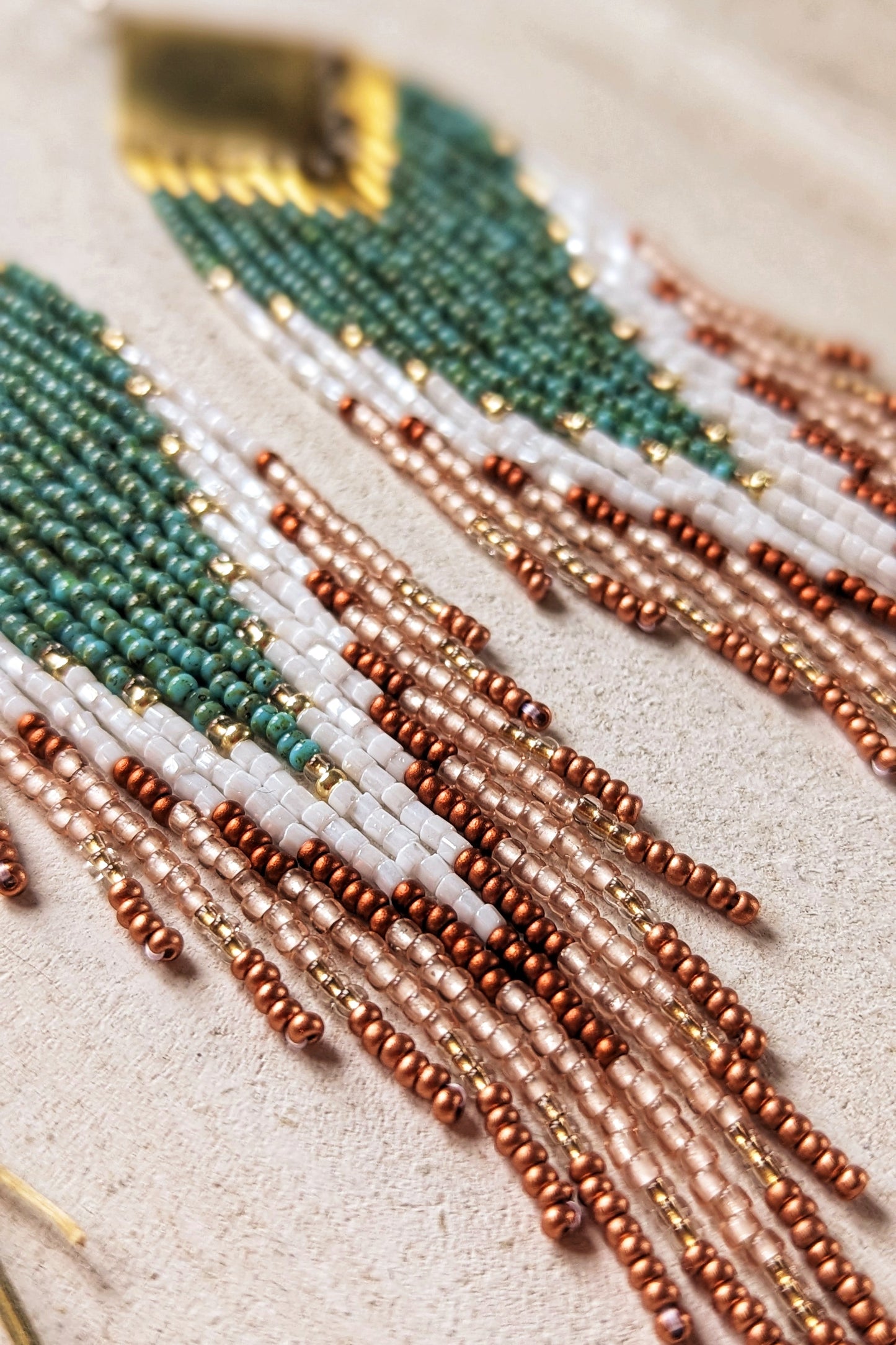 Close-up of handcrafted Moon & Milk earrings with turquoise, white, and crimson beads arranged in a chevron pattern, hanging from brass diamond-shaped findings.