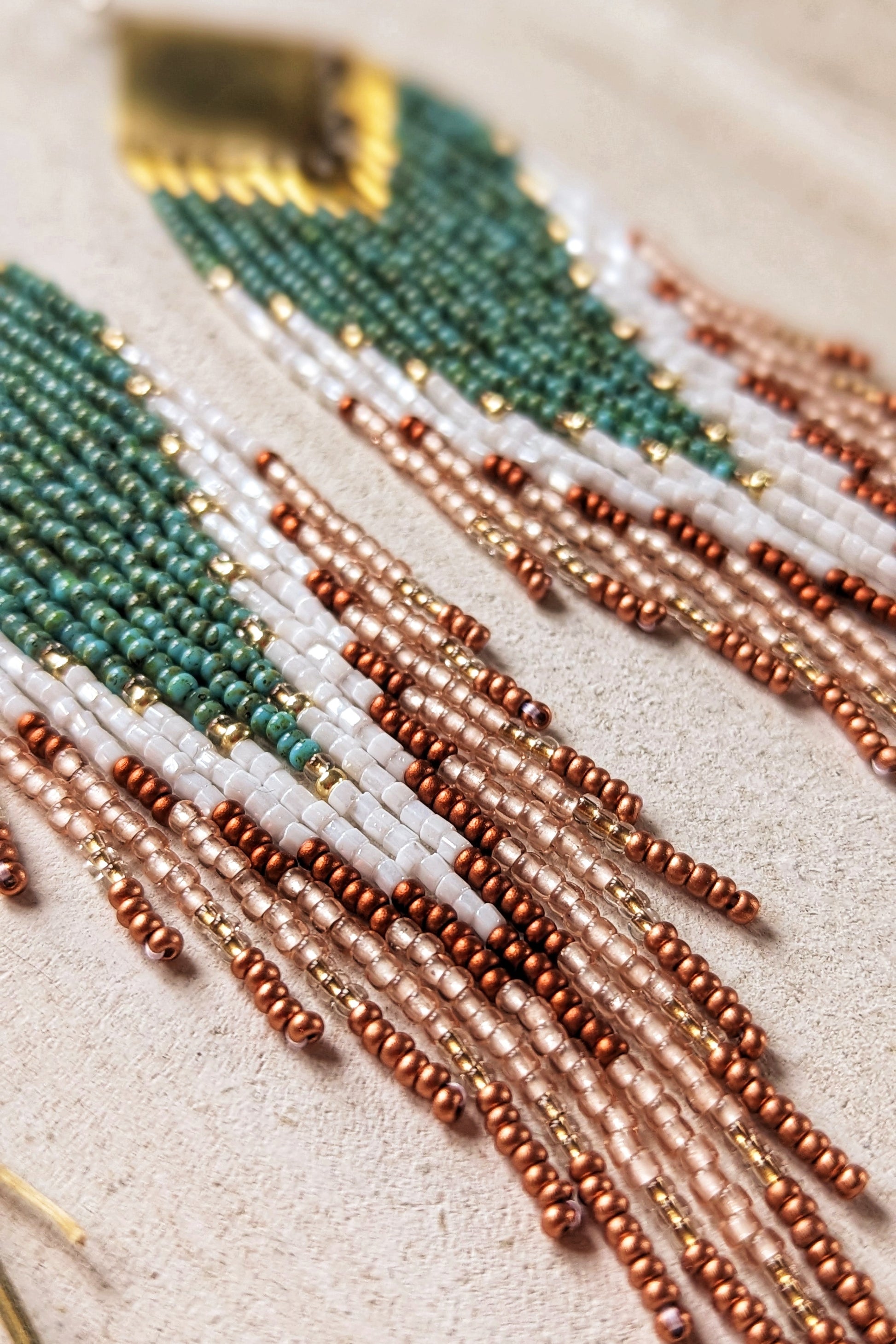 Close-up of handcrafted Moon & Milk earrings with turquoise, white, and crimson beads arranged in a chevron pattern, hanging from brass diamond-shaped findings.