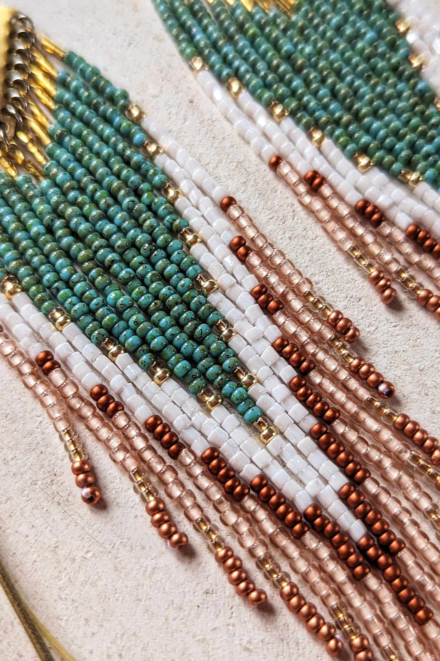Close-up of handcrafted Moon & Milk earrings with turquoise, white, and crimson beads arranged in a chevron pattern, hanging from brass diamond-shaped findings.