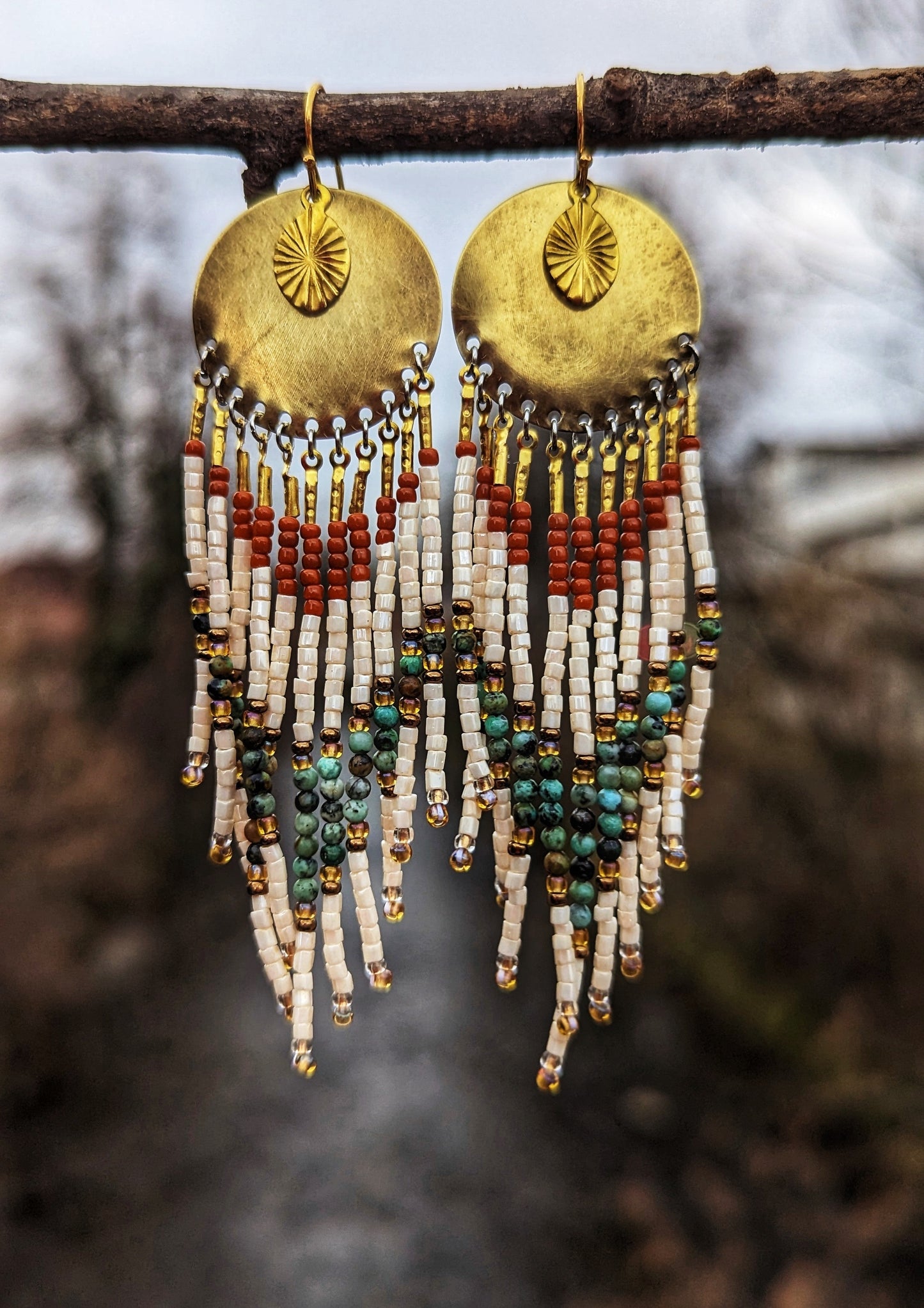 Moon & Milk - Gold dangling earrings with brass circular charms featuring sunburst designs. Hanging below are strands of white, bronze, burnt orange, and turquoise stones. Photographed outdoors on a branch.