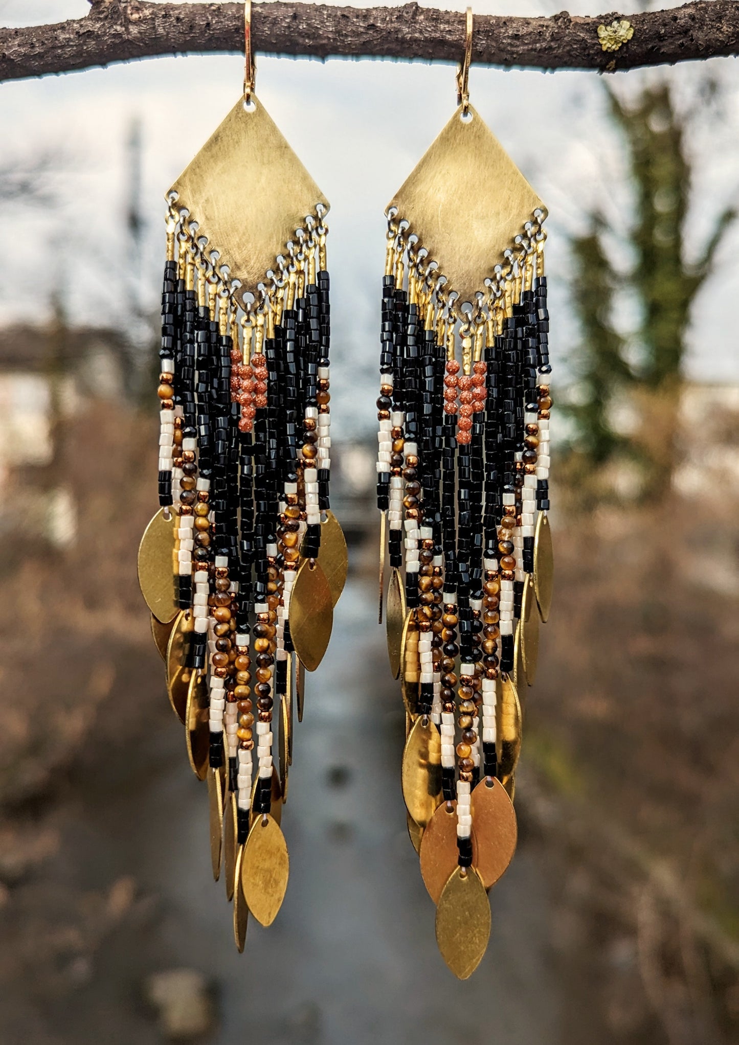 Moon & Milk - Long seed bead earrings with black, gold, white, brown beads, and tiger eye stones, in a geometric pattern, featuring gold brass diamond-shaped tops and hanging gold leaf accents. Photographed in a forest.