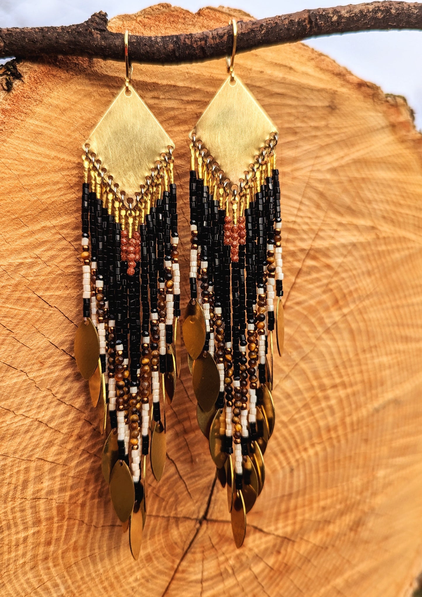 Moon & Milk - Long seed bead earrings with black, gold, white, brown beads, and tiger eye stones, in a geometric pattern, featuring gold brass diamond-shaped tops and hanging gold leaf accents. Photographed on a wooden background.
