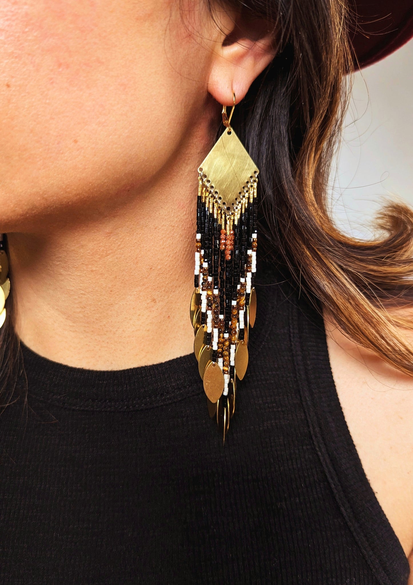 Moon & Milk - Long seed bead earrings with black, gold, white, brown beads, and tiger eye stones, in a geometric pattern, featuring gold brass diamond-shaped tops and hanging gold leaf accents. Photographed on a woman with brown hair.