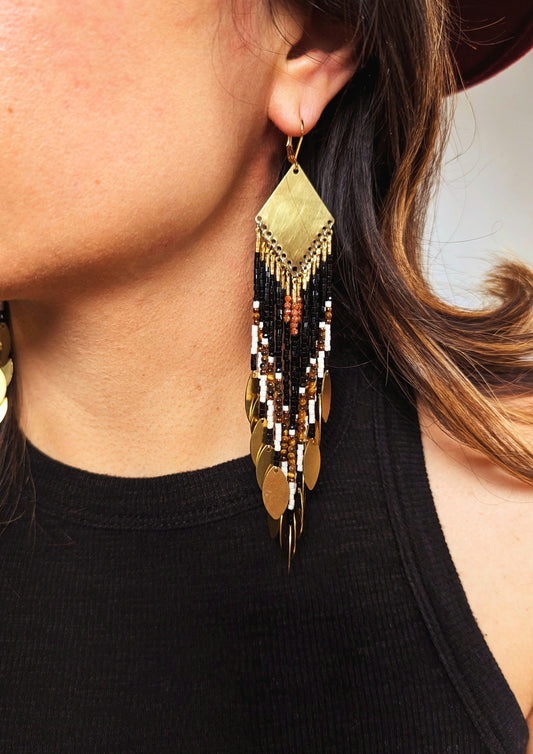 Moon & Milk - Long seed bead earrings with black, gold, white, brown beads, and tiger eye stones, in a geometric pattern, featuring gold brass diamond-shaped tops and hanging gold leaf accents. Photographed on a woman with brown hair.