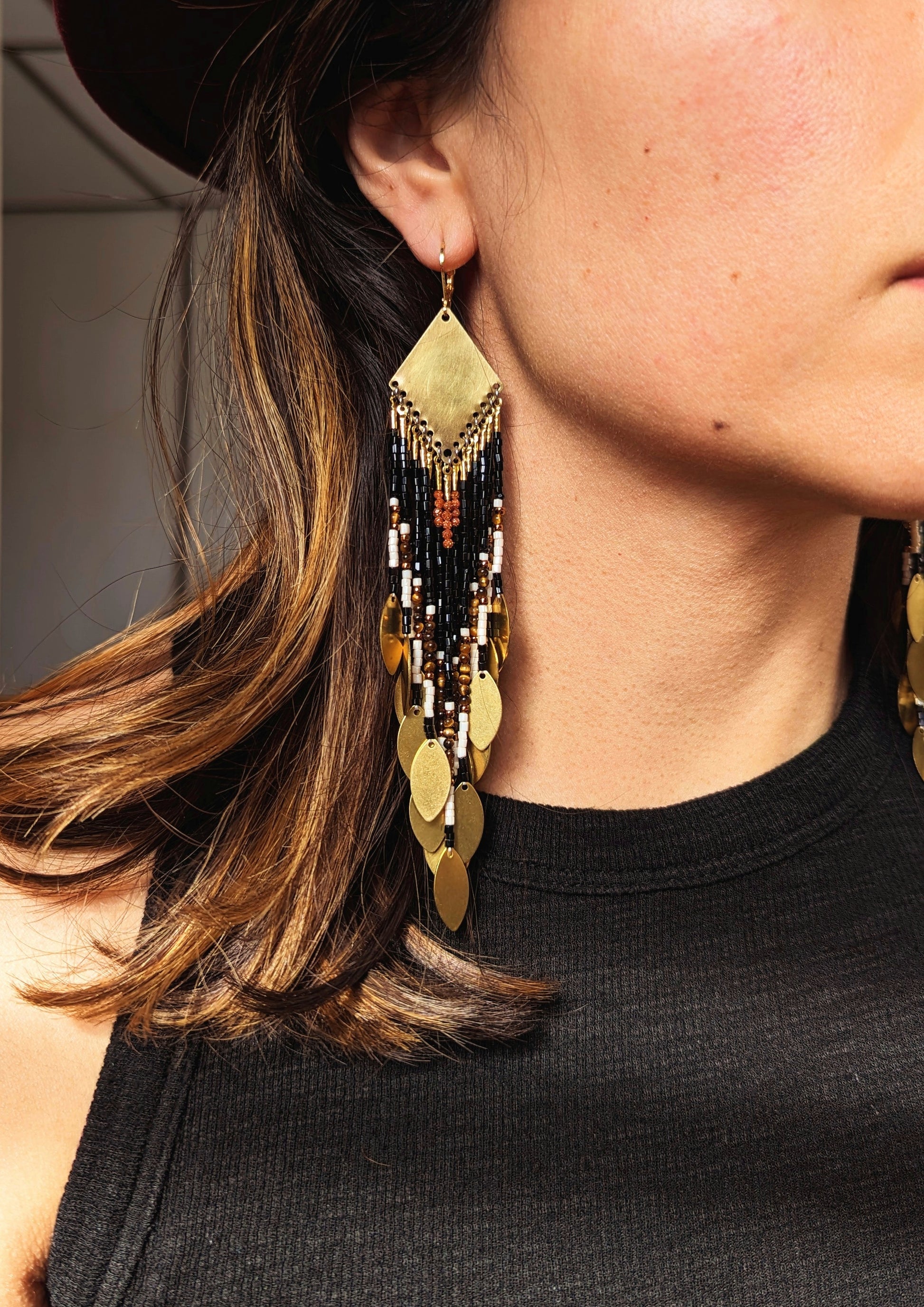 Moon & Milk - Long seed bead earrings with black, gold, white, brown beads, and tiger eye stones, in a geometric pattern, featuring gold brass diamond-shaped tops and hanging gold leaf accents. Photographed on a woman with brown hair.