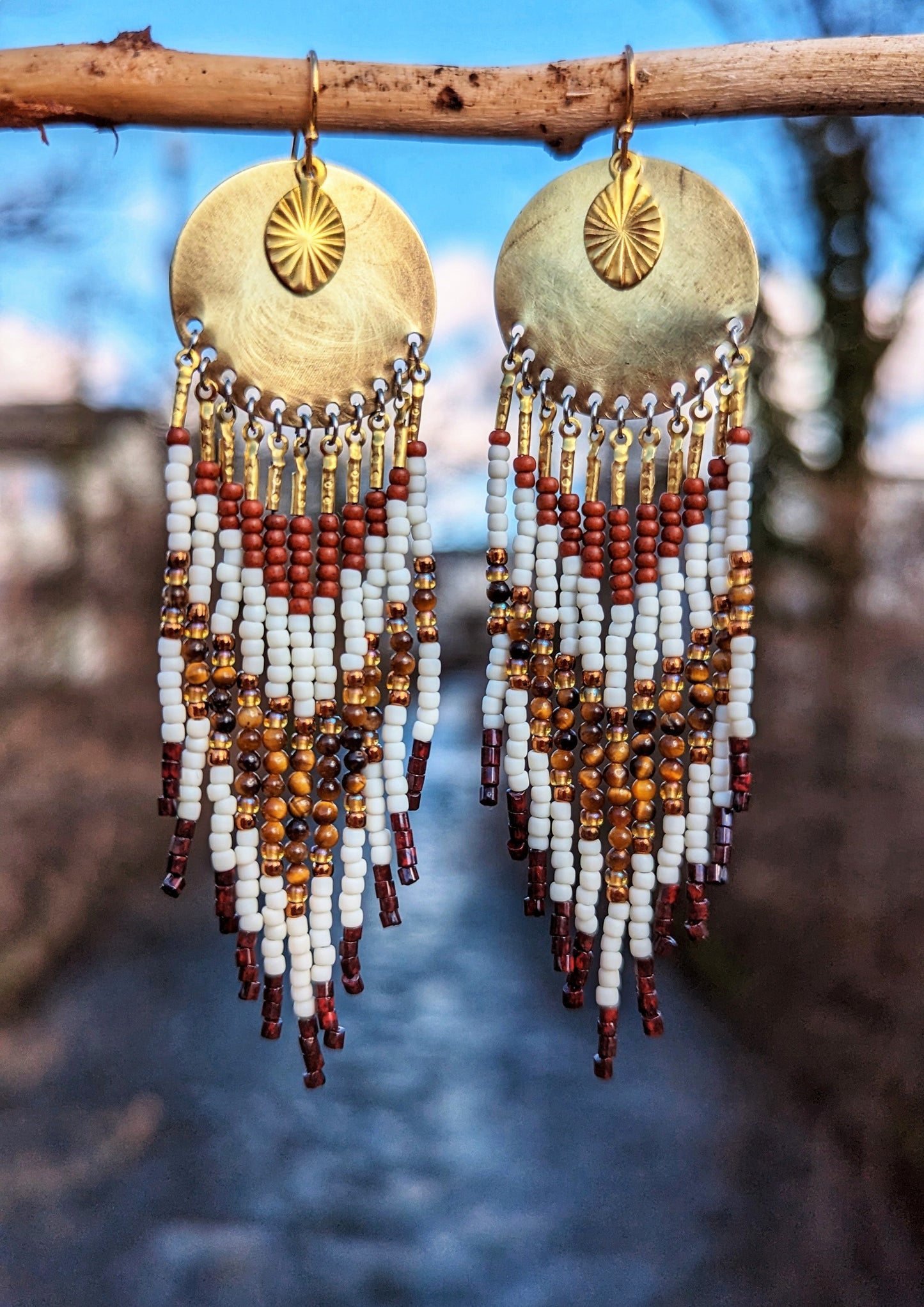 Moon & Milk handcrafted earrings with ivory, amber, and rust beadwork, brass discs, and sun charms hanging from a branch with a soft blue sky background.