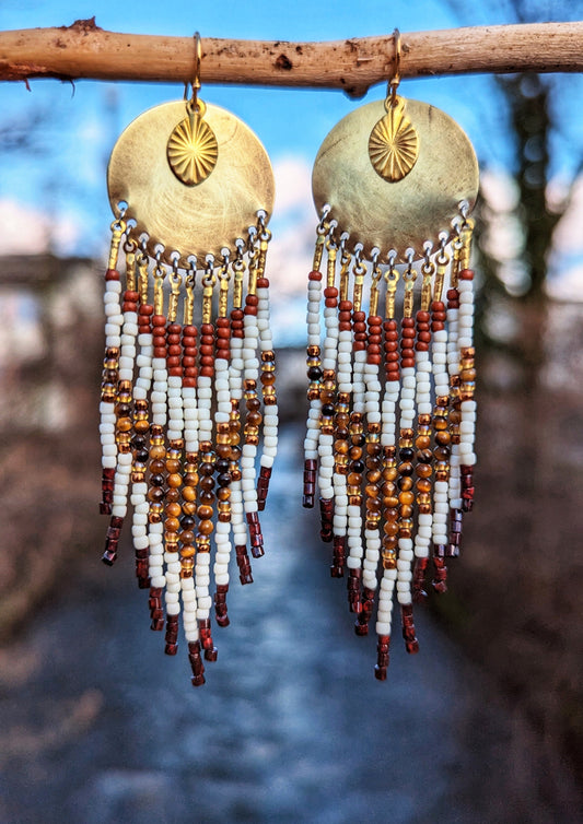 Moon & Milk handcrafted earrings with ivory, amber, and rust beadwork, brass discs, and sun charms hanging from a branch with a soft blue sky background.