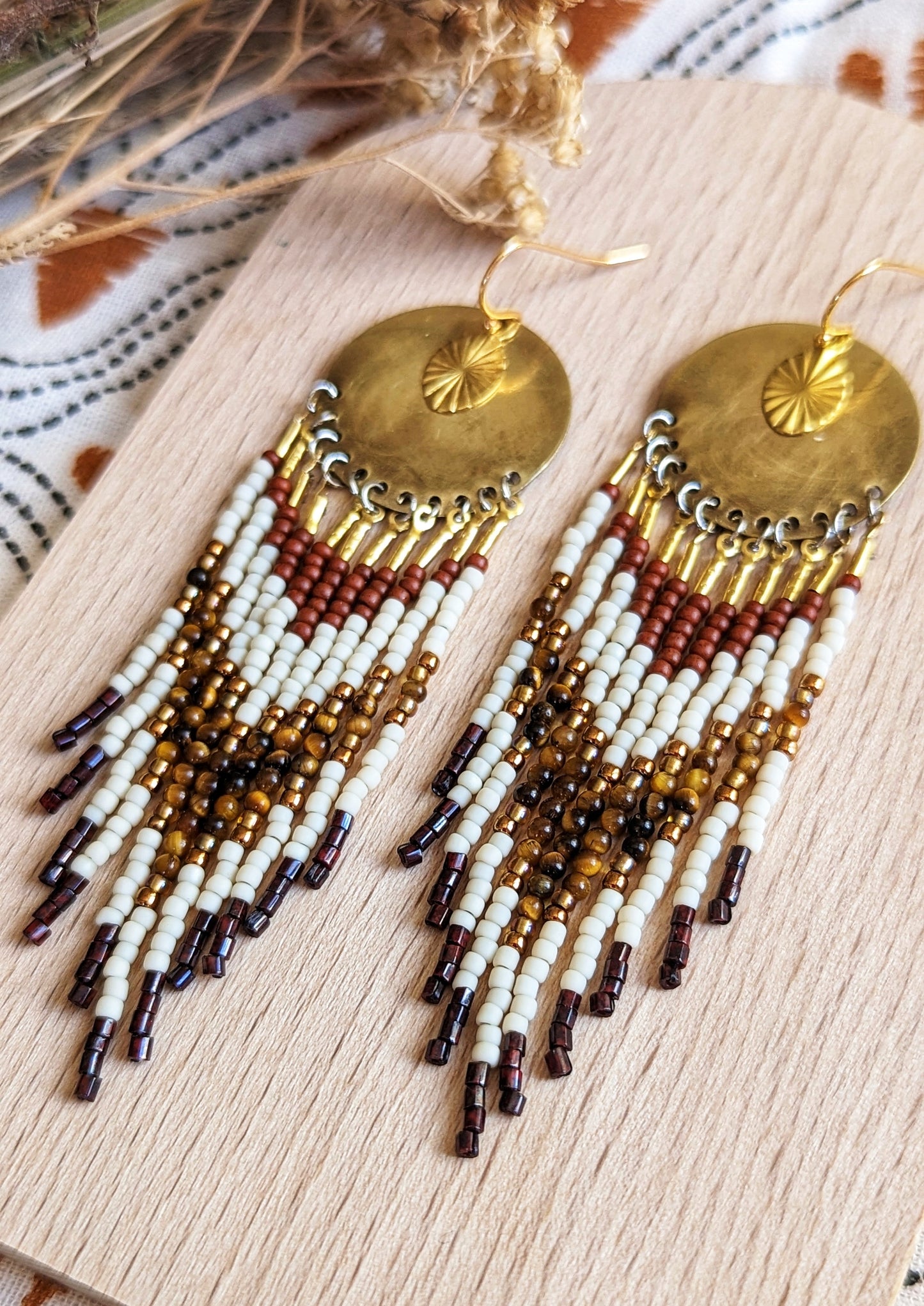 Moon & Milk handcrafted earrings with ivory, amber, and rust beadwork, brass discs, and sun charms laid flat on a wooden coaster.