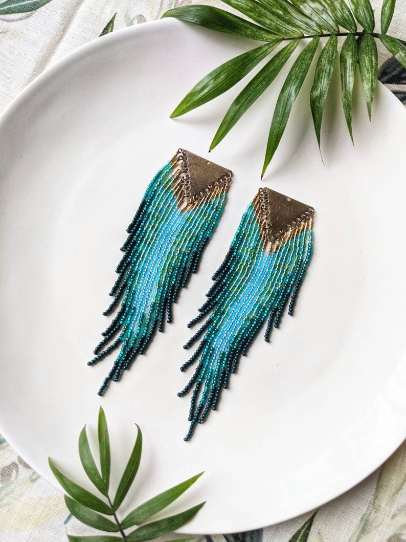 Pair of seed bead earrings with a triangle brass top, featuring cascading strands in shades of blue and green, displayed on a white plate surrounded by green leaves.