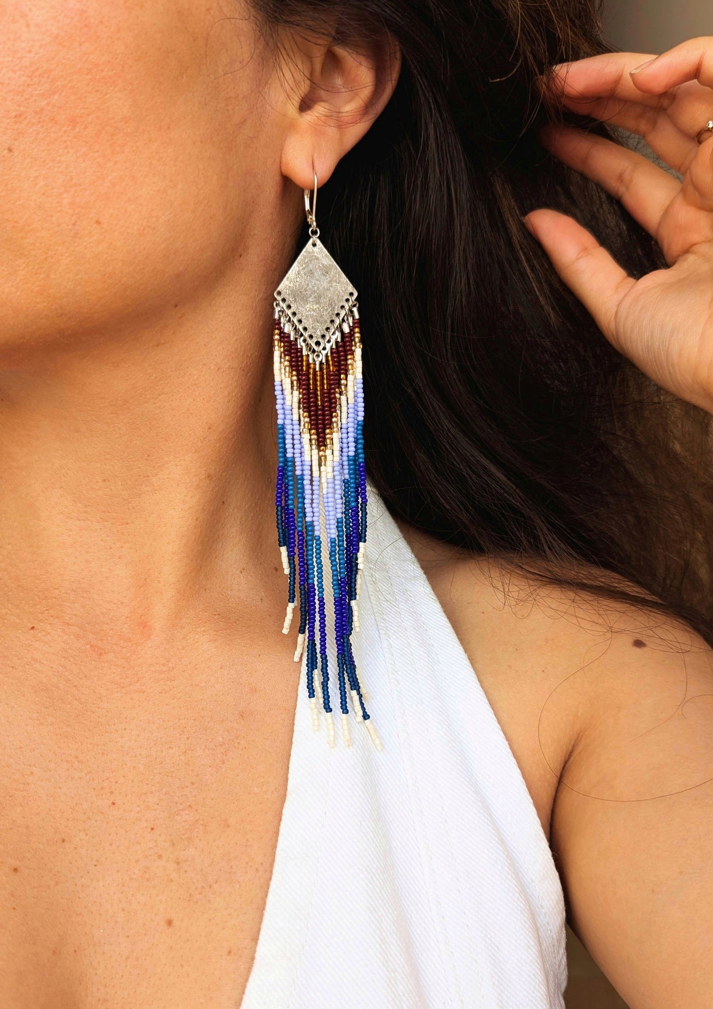 Close-up of a woman wearing long beaded earrings with a silver top, featuring a blue, white, and brown gradient pattern, paired with a white outfit. - Moon & Milk