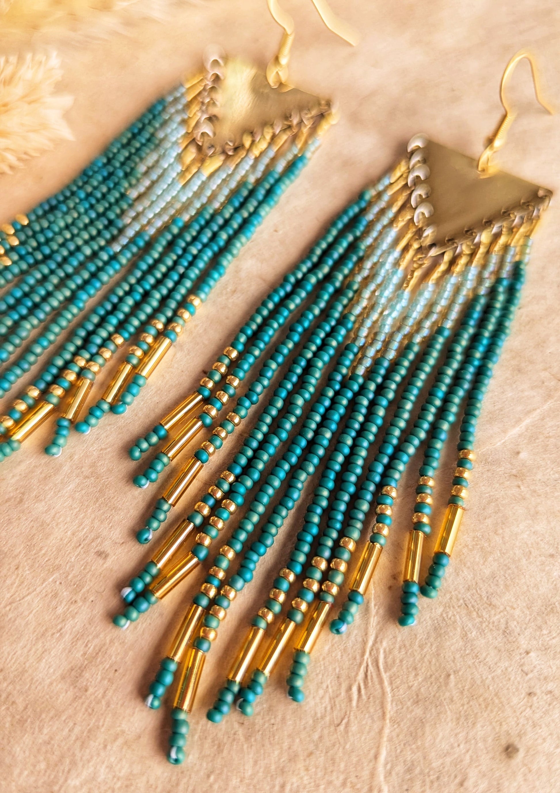 Moon & Milk - Long seed bead earrings with blue-green, gold, and light blue beads in a chevron pattern. Gold brass triangular tops. Photographed on a beige background with dried grasses.