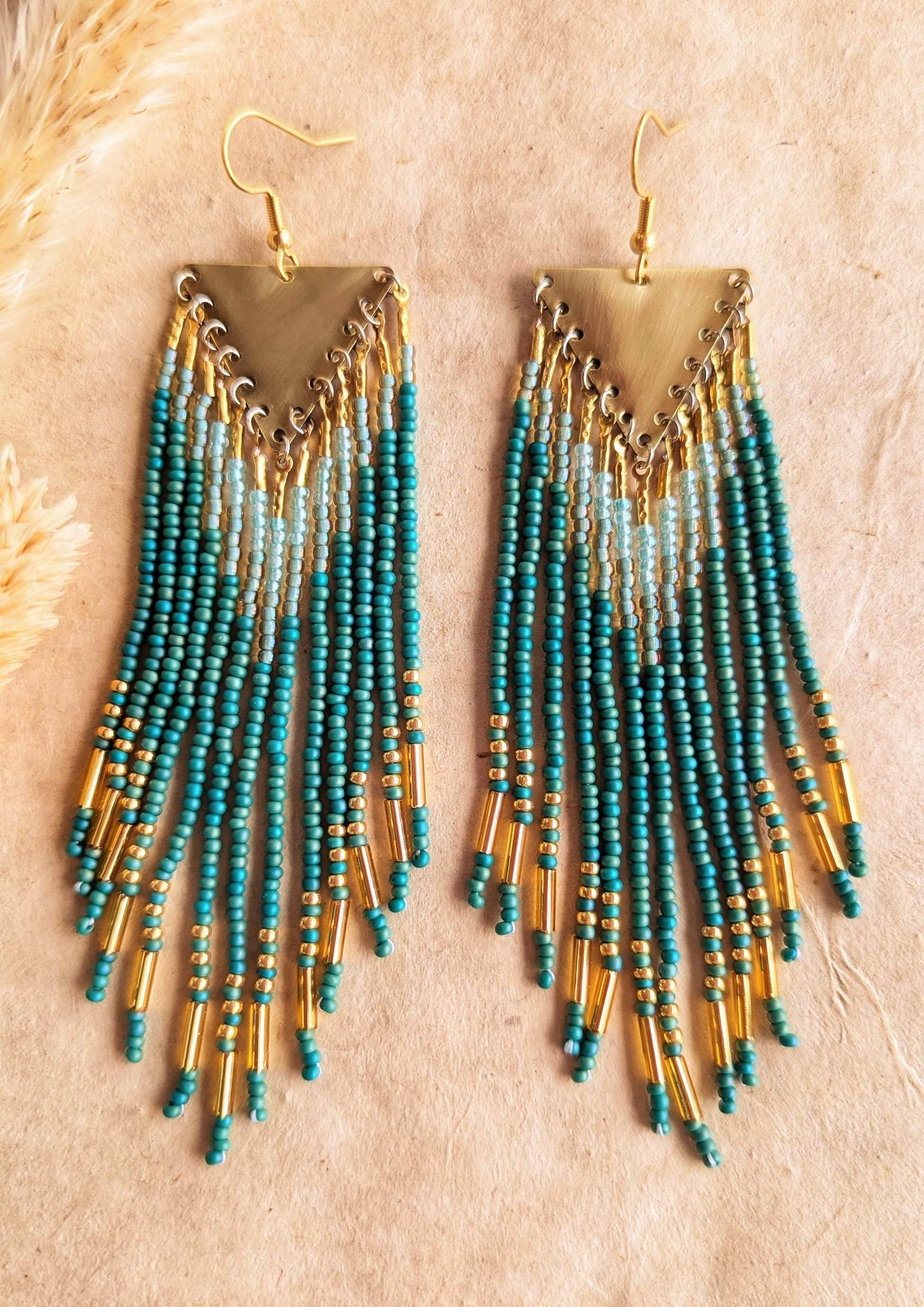 Moon & Milk - Long seed bead earrings with blue-green, gold, and light blue beads in a chevron pattern. Gold brass triangular tops. Photographed on a beige background with dried grasses.