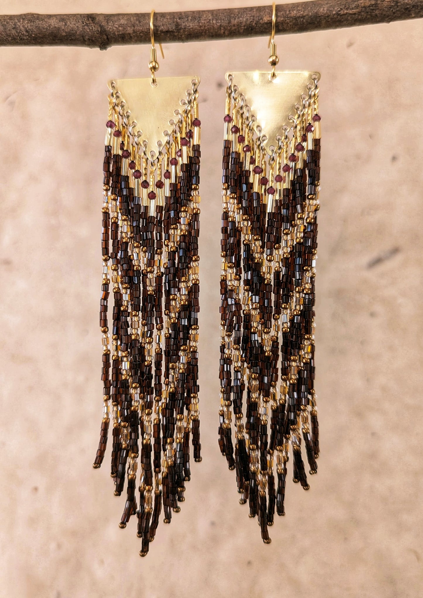 Moon & Milk - Long seed bead earrings with brown, gold, and bronze beads in a chevron pattern. Gold brass triangular tops. Photographed on a beige background hanging from a bamboo stick.