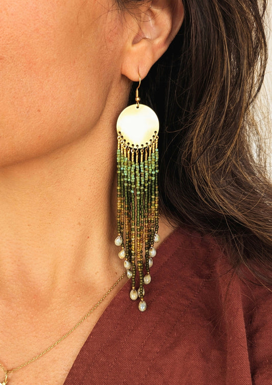 Close-up of a woman wearing long beaded green earrings with brass disks and glass teardrops, styled with a burgundy top.