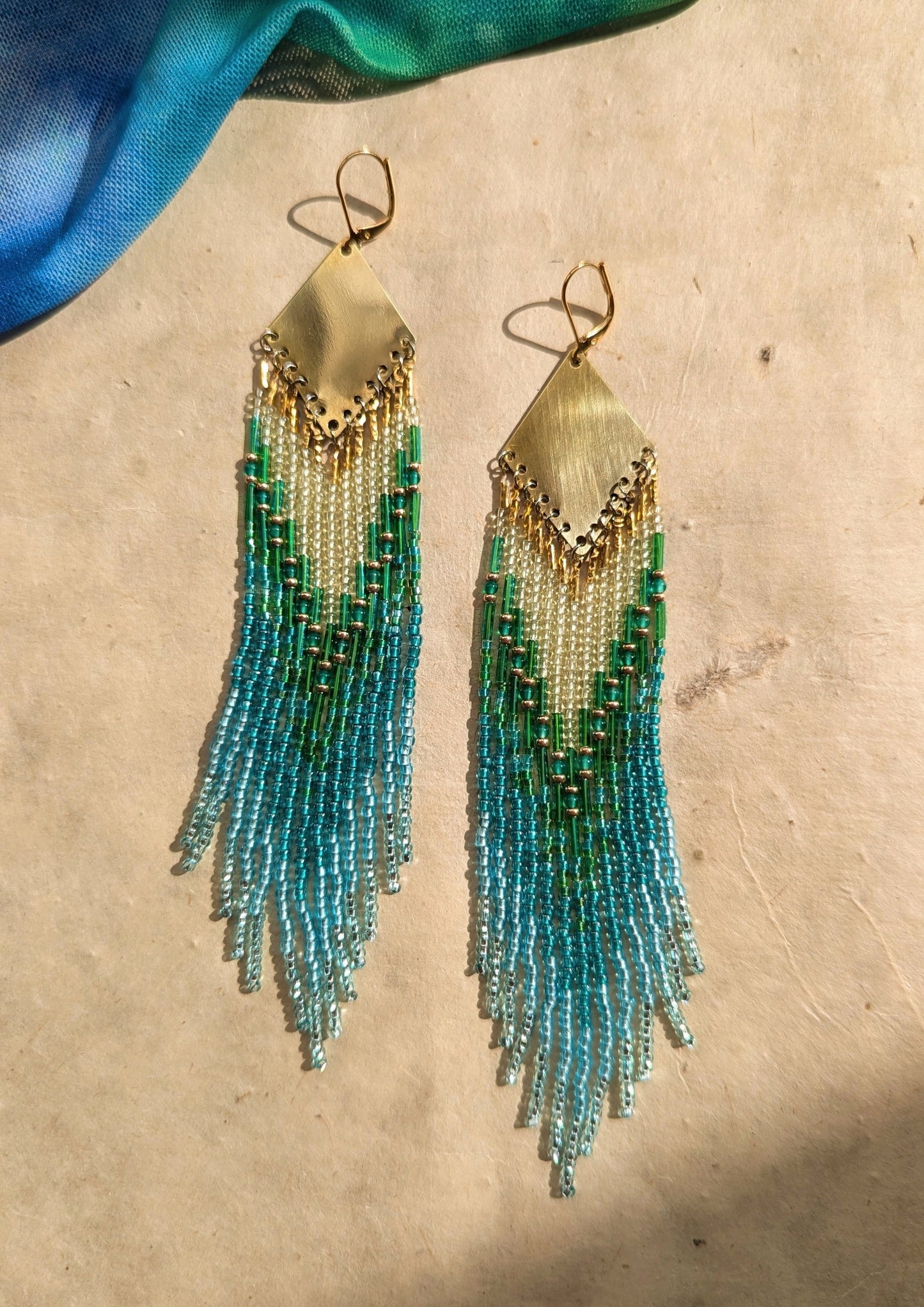 Moon & Milk - Handcrafted seed bead earrings in oceanic shades of blue and green, elegantly dangling from a brass diamond-shaped plate, beautifully contrasted against a light background with a touch of blue fabric.