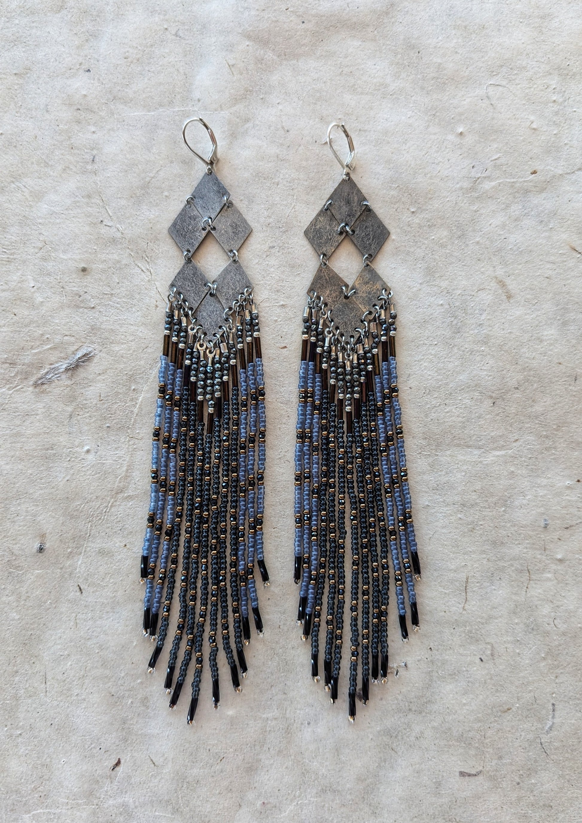 Moon & Milk - Handmade beaded earrings featuring a unique geometric design with metallic gray diamonds and cascading strands of black, blue, and bronze beads, displayed on a natural textured background.