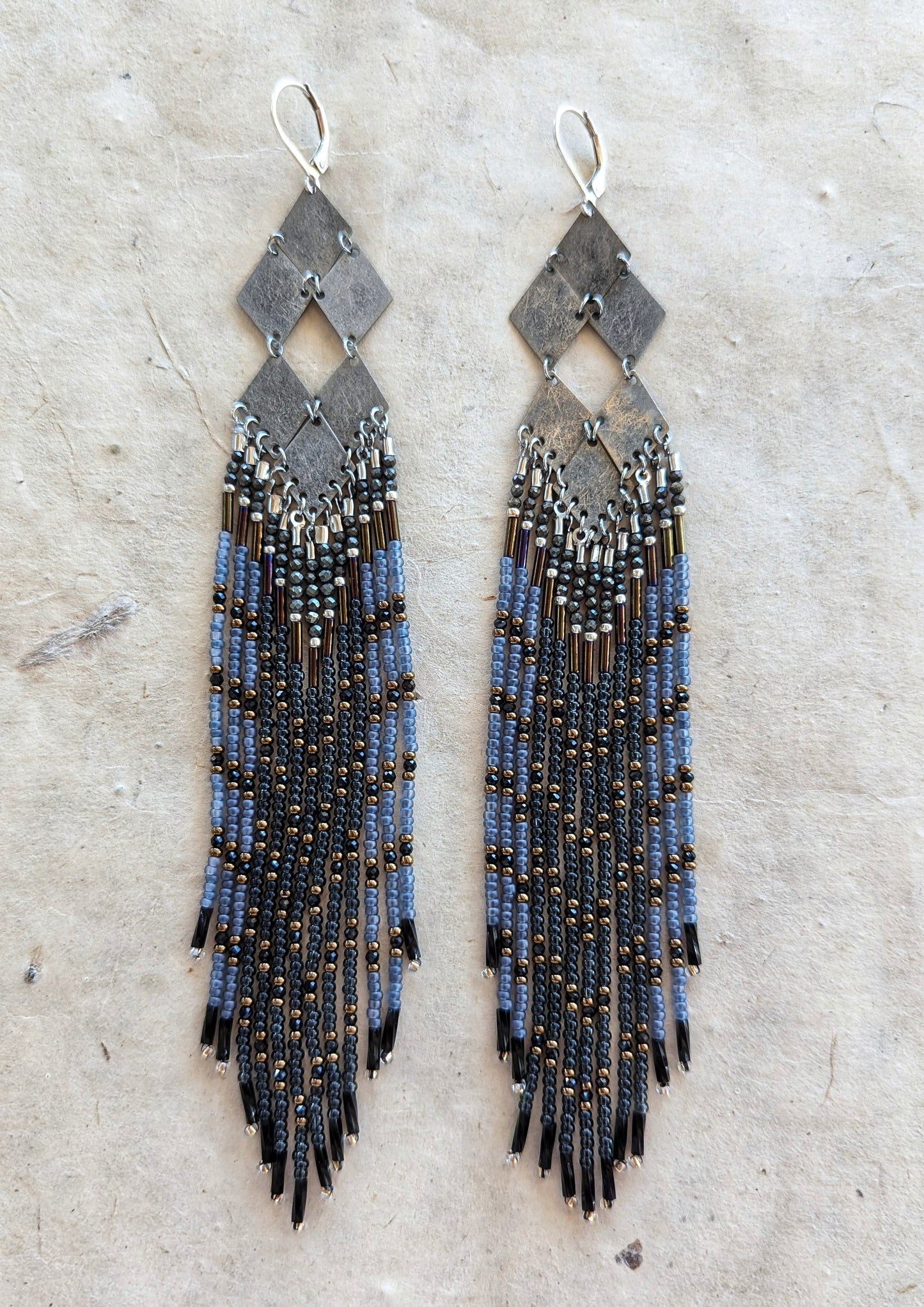 Moon & Milk - Handmade beaded earrings featuring a unique geometric design with metallic gray diamonds and cascading strands of black, blue, and bronze beads, displayed on a natural textured background.