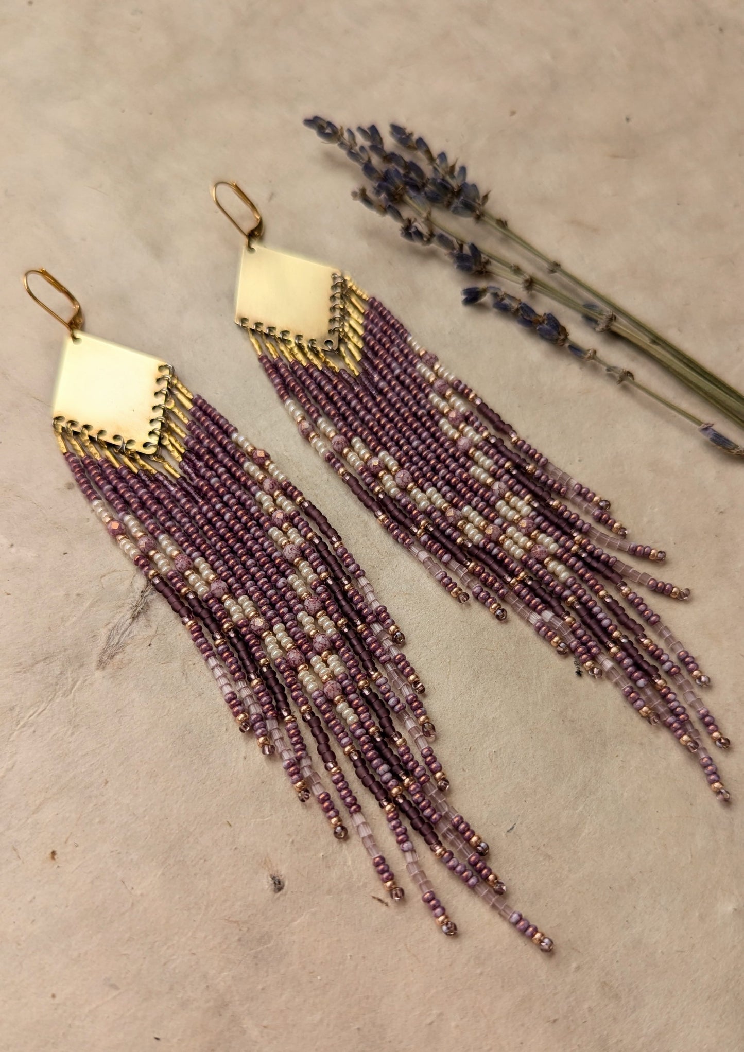 Moon & Milk - beaded earrings featuring purple and cream strands cascading from a gold diamond-shaped brass pendant, displayed on a neutral background with sprigs of lavender.