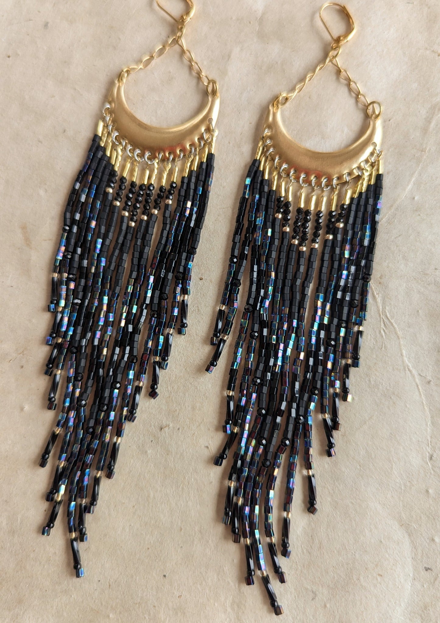 Moon & Milk - Gold crescent-shaped earrings with cascading black iridescent bead strands, displayed on a neutral background.