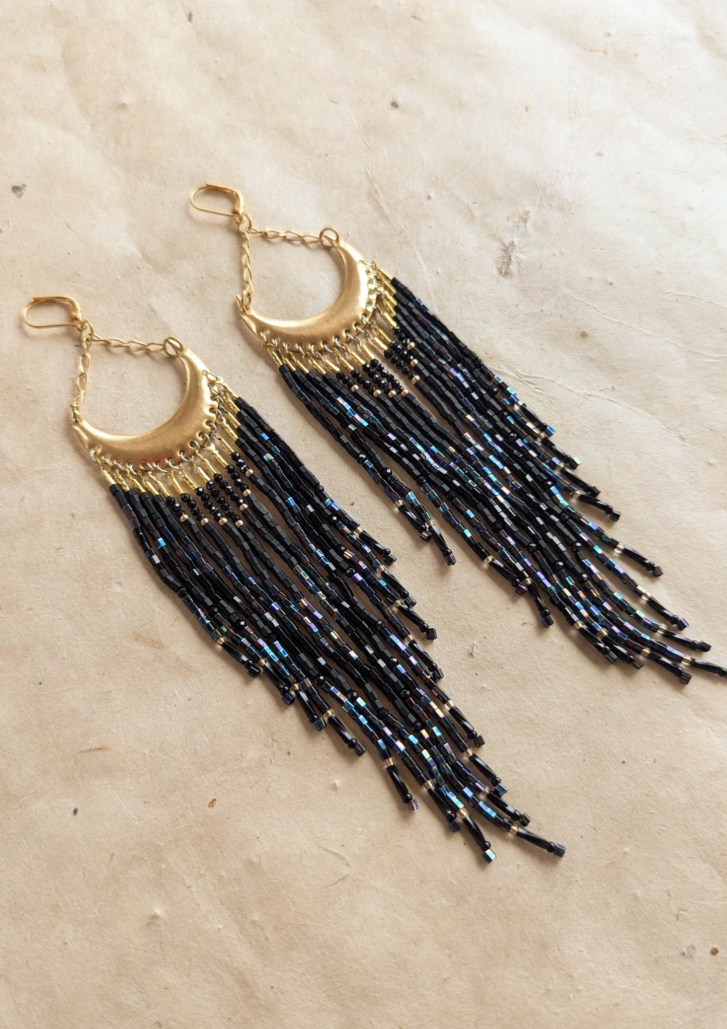 Moon & Milk - Gold crescent-shaped earrings with cascading black iridescent bead strands, displayed on a neutral background.