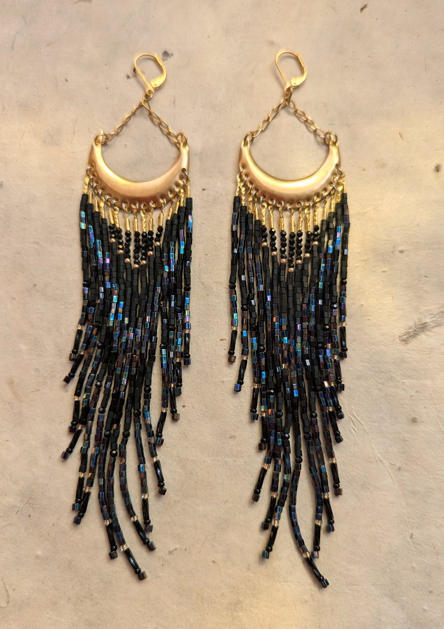 Moon & Milk - Gold crescent-shaped earrings with cascading black iridescent bead strands, displayed on a neutral background.