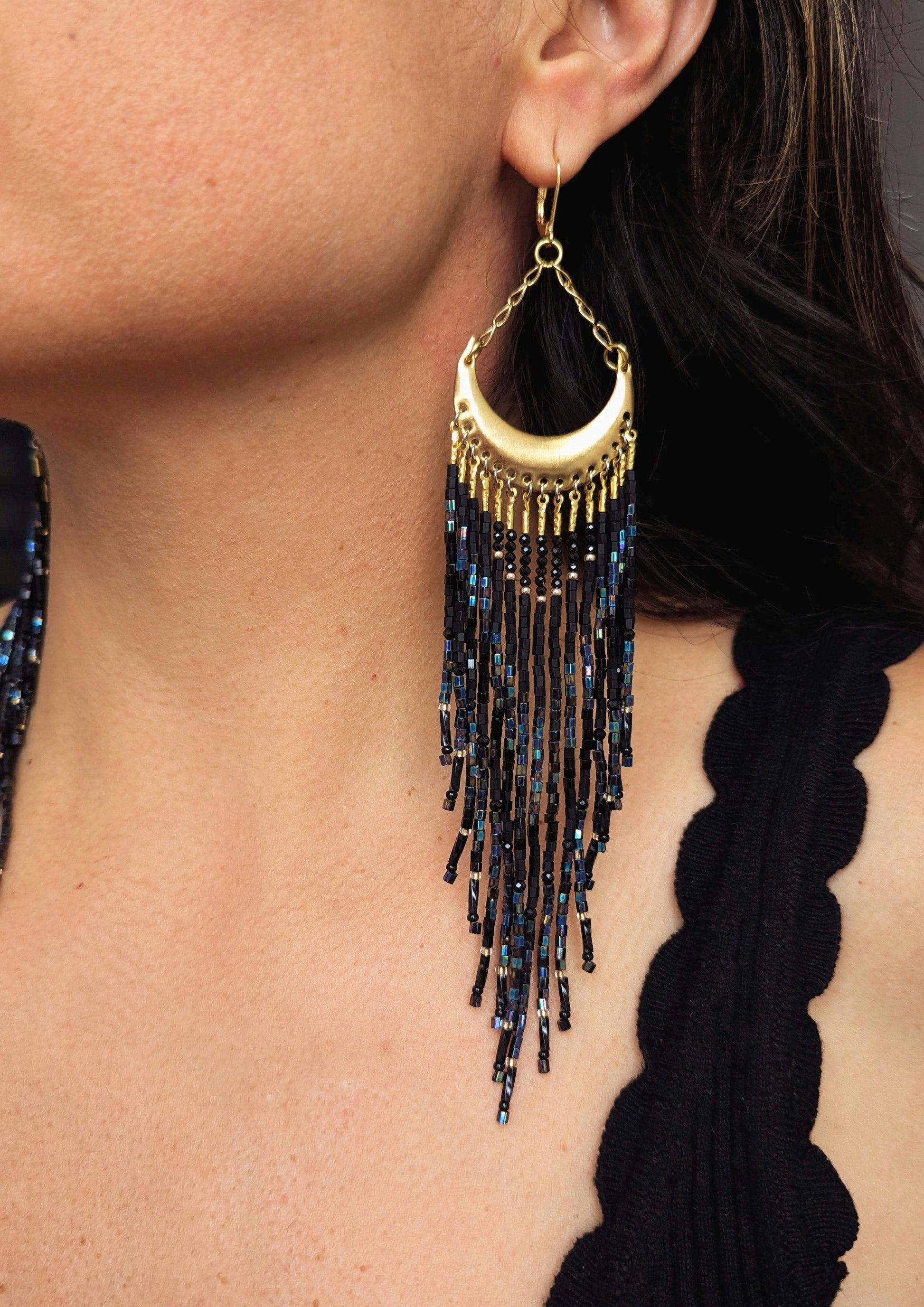 Moon & Milk - Model wearing a striking crescent-shaped gold earring adorned with shimmering black and blue seed bead fringes, creating a bold, elegant statement.