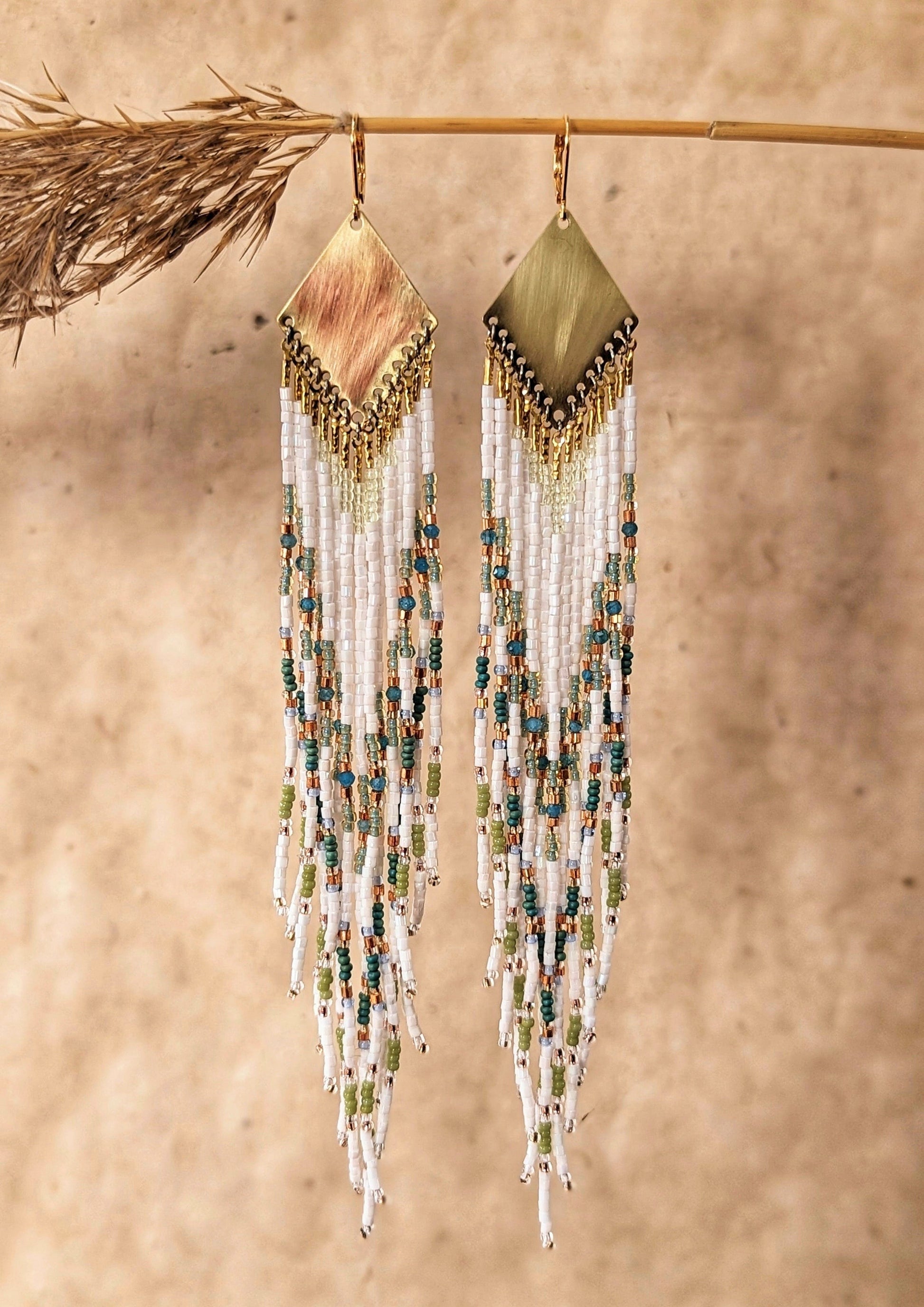 Moon & Milk - Long seed beaded earrings with white, green, blue beads, and apatite stones in a chevron pattern. Gold diamond-shaped brass charms at the top. Photographed on a beige background