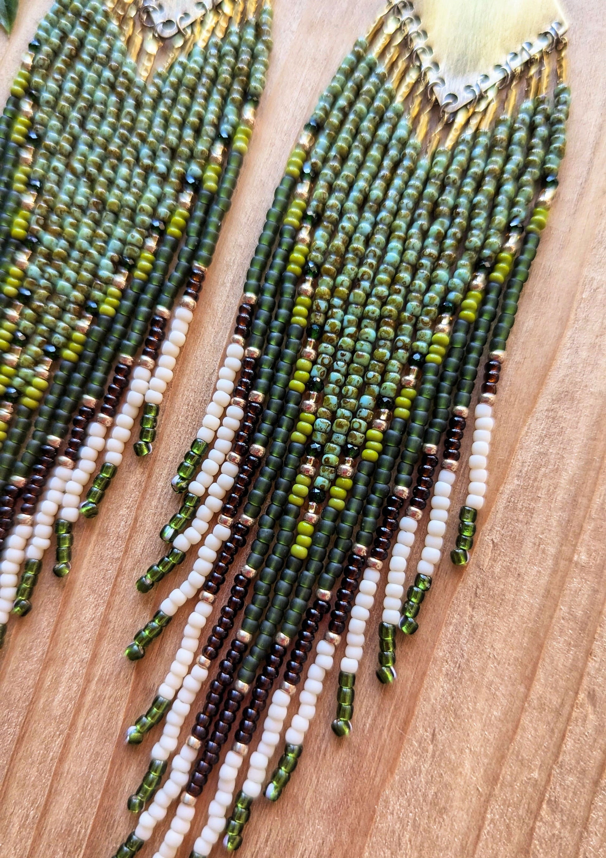 Pair of handmade beaded earrings with brass tops, featuring green, brown, and white beads, laid on a wooden surface next to vibrant green leaves. - Moon & Milk 