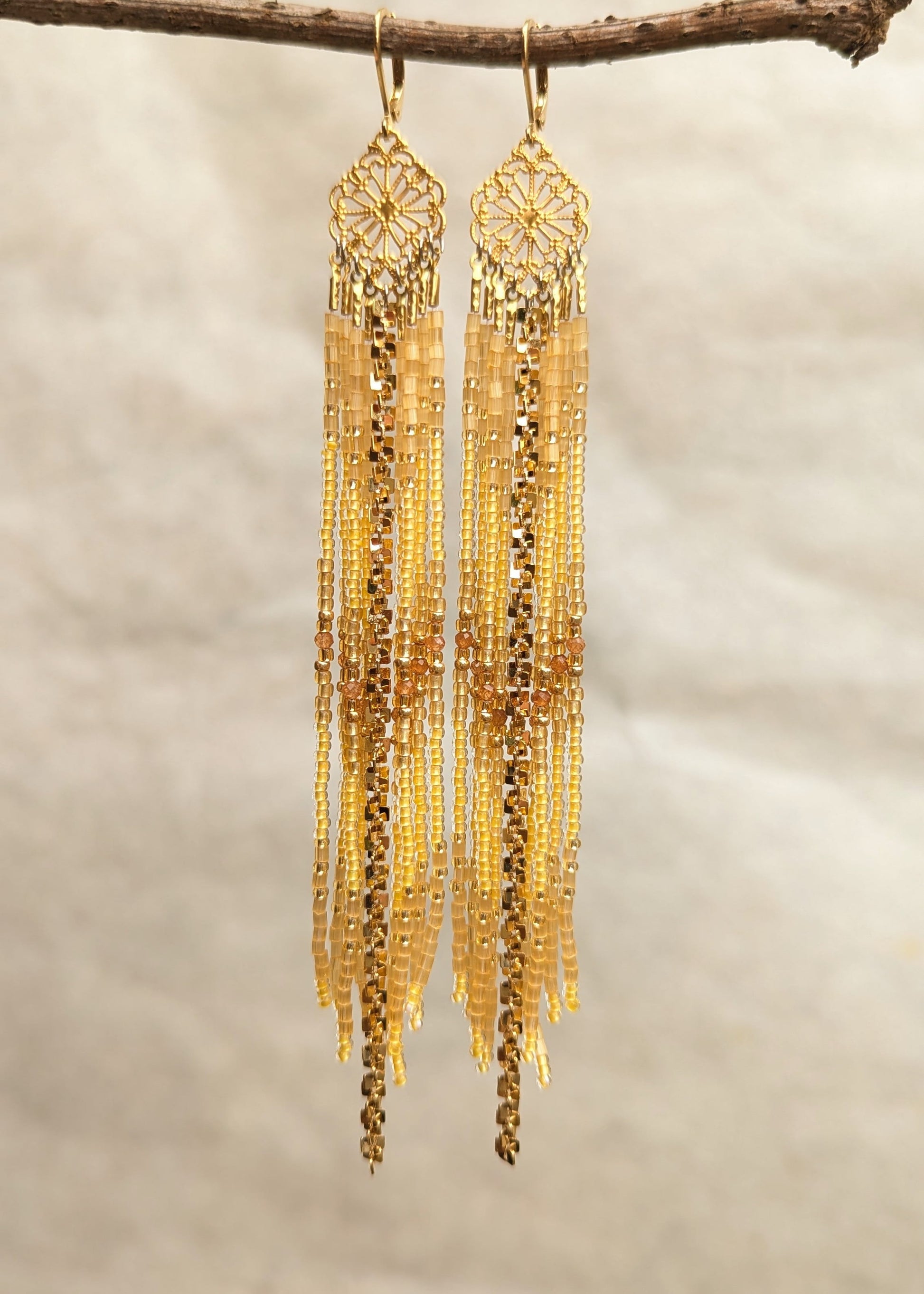 A pair of long gold-toned dangle beaded earrings with intricate filigree tops and layered chain fringe, hanging from a small branch against a soft beige textured background.