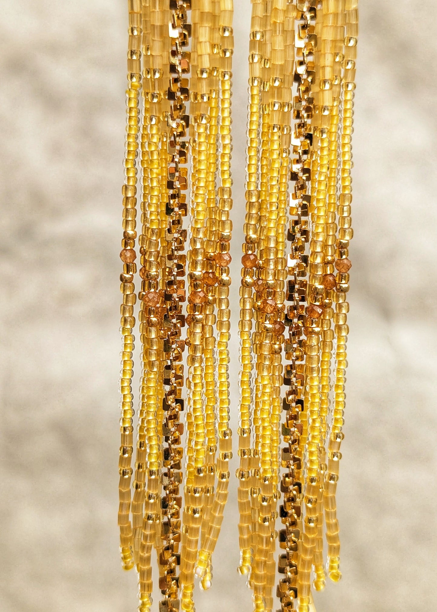 Seed bead earrings in shades of yellow, gold, and amber, hanging vertically against a soft, out-of-focus neutral background.