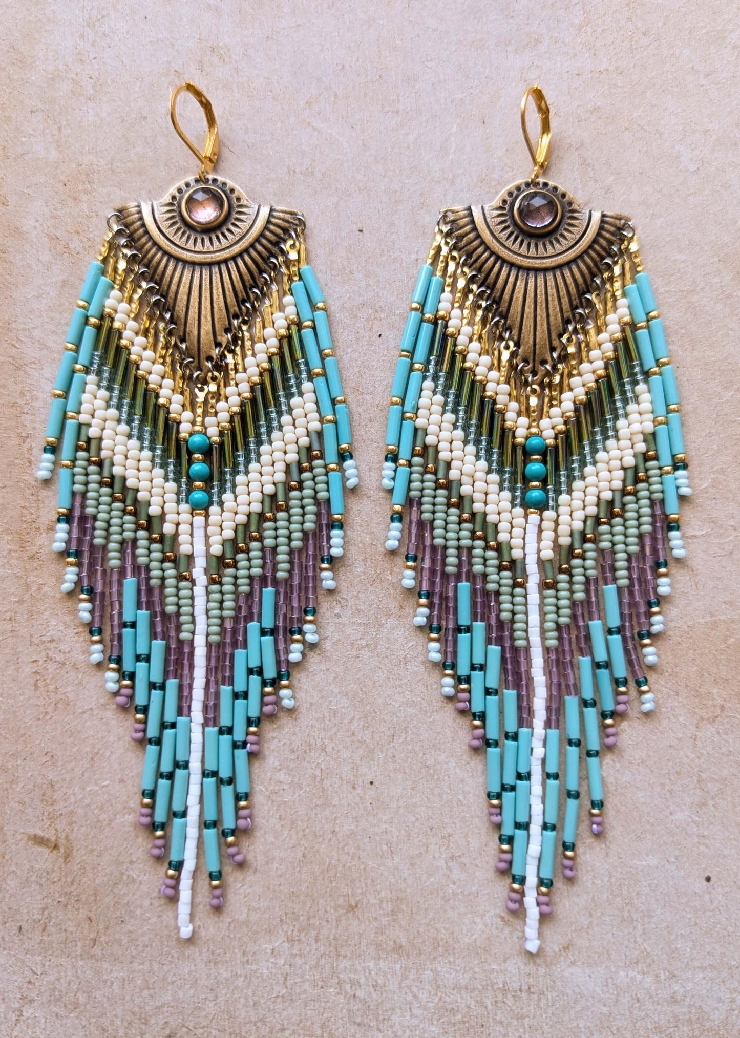A pair of bohemian beaded fringe dangle earrings with turquoise, cream, and purple beads on a textured beige background by Moon & Milk. 