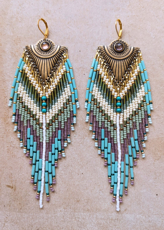 A pair of bohemian beaded fringe dangle earrings with turquoise, cream, and purple beads on a textured beige background by Moon & Milk. 