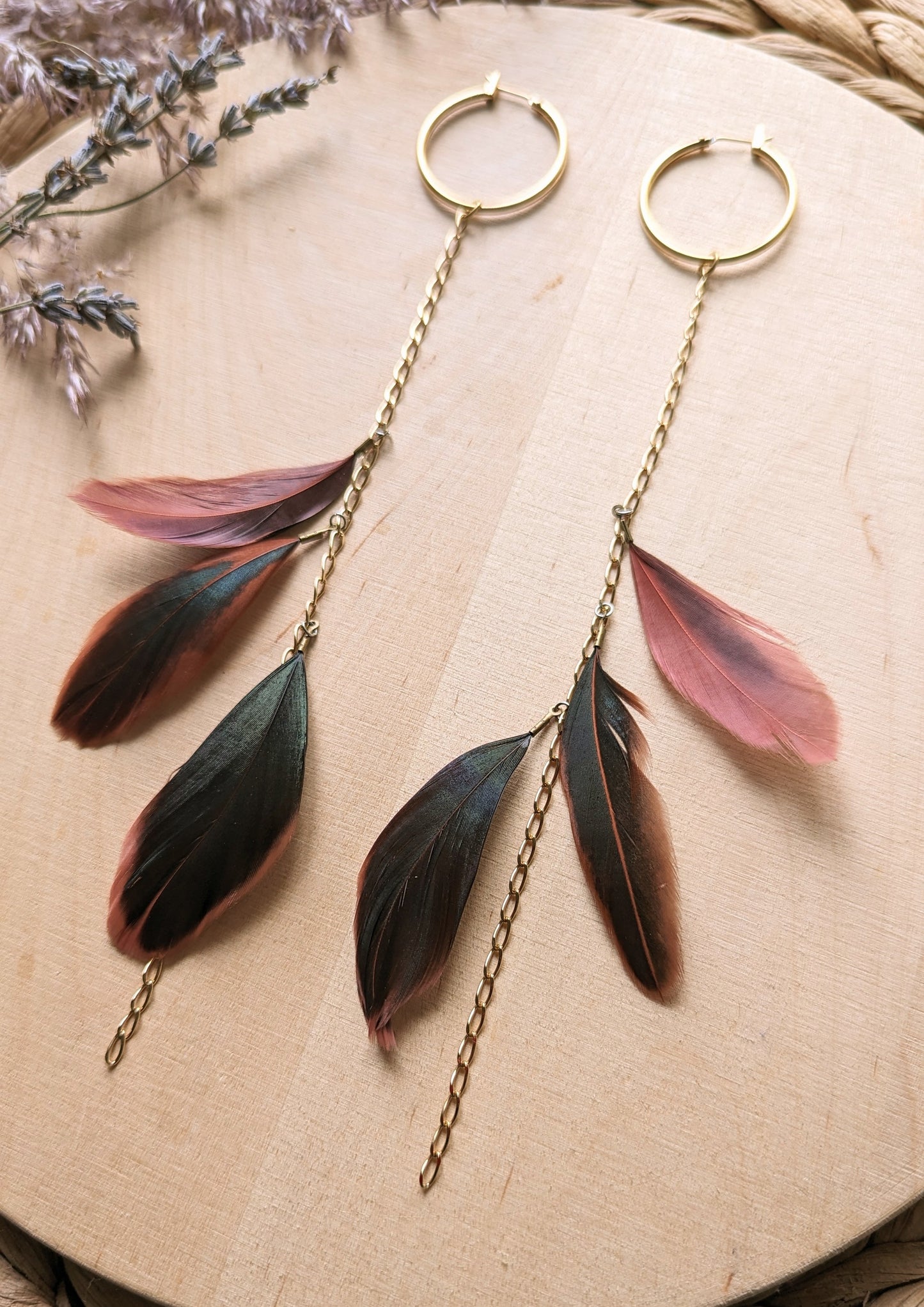 Gold hoop earrings with delicate chain drops, featuring layered feathers in deep brown and muted pink tones, styled on a wooden surface with lavender sprigs.