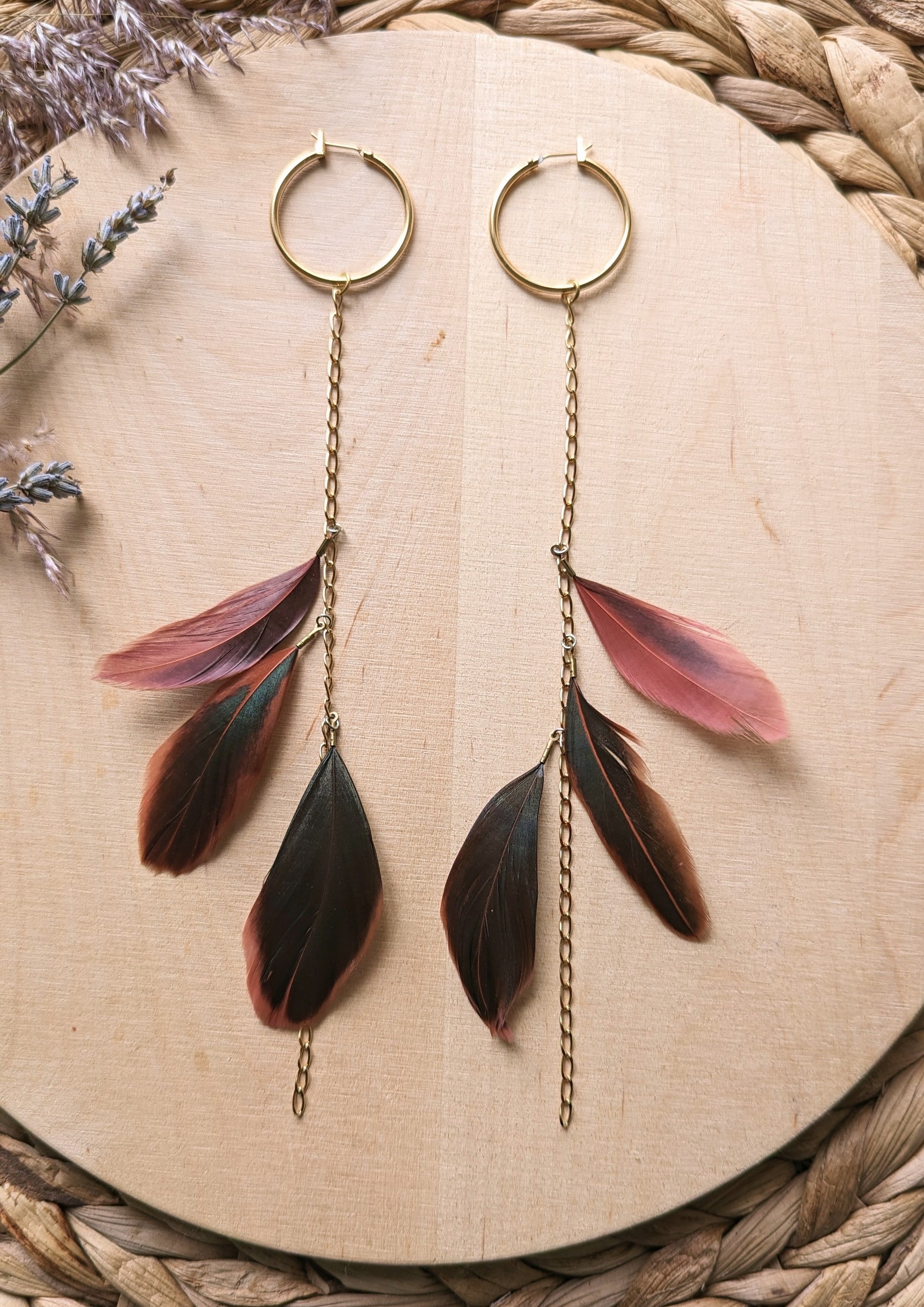 Gold hoop earrings with delicate chain drops, featuring layered feathers in deep brown and muted pink tones, styled on a wooden surface with lavender sprigs.