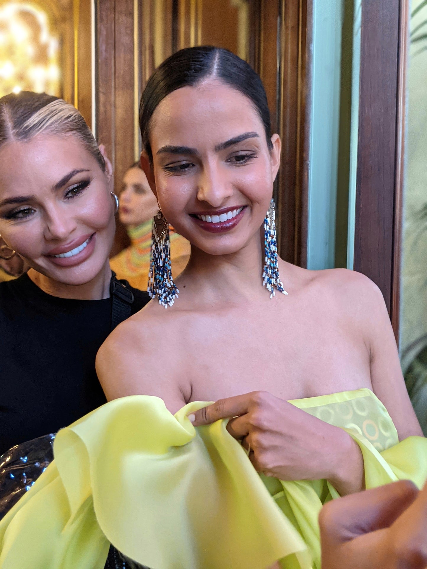 A model wearing bold, handmade beaded fringe earrings with intricate patterns, paired with a strapless lime green dress, walking in a brightly lit venue.