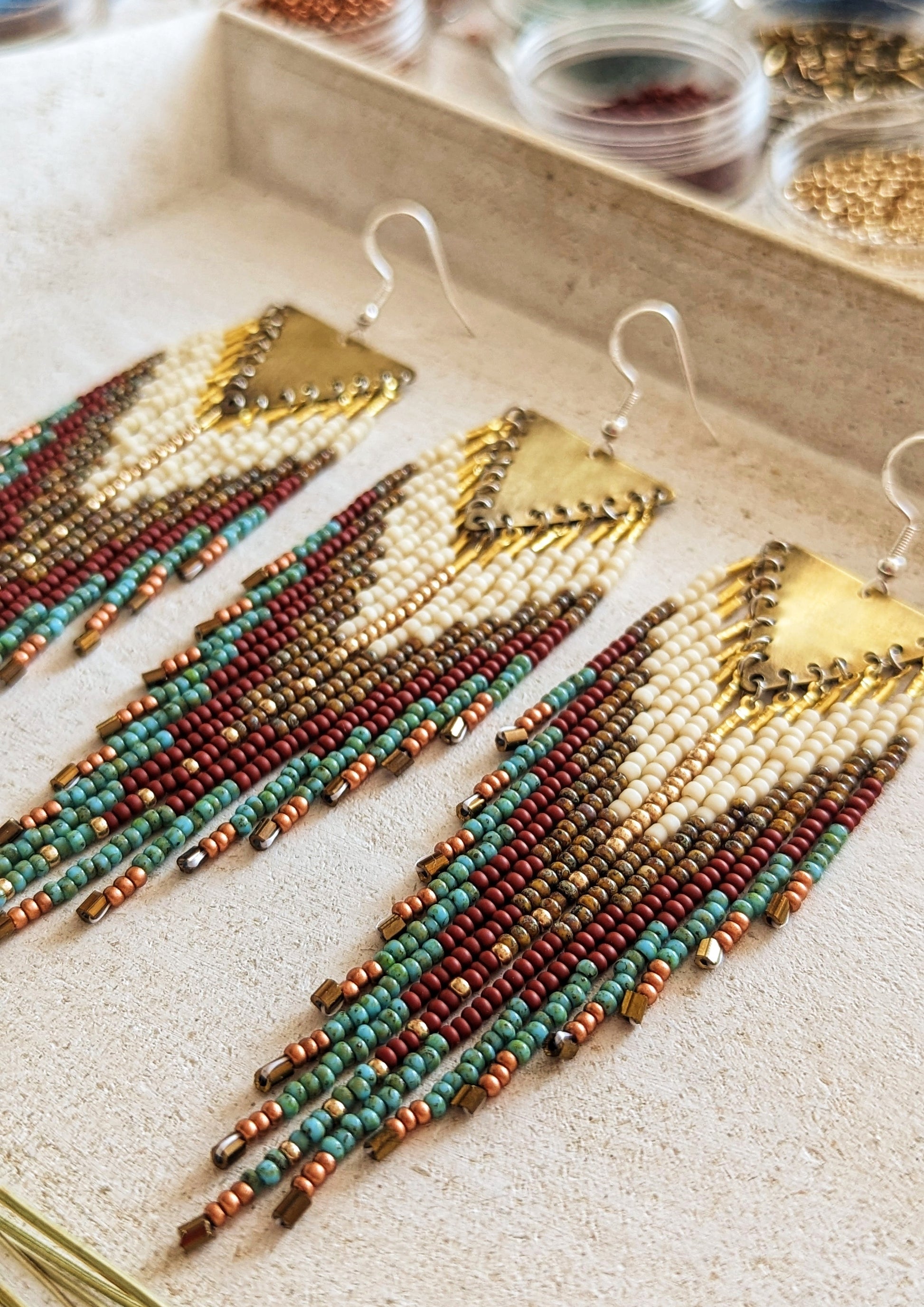 Moon & Milk - Elegant bohemian-style earrings with intricate beadwork featuring a combination of ivory, maroon, turquoise, and bronze beads, complemented by a brass chevron pendant. Perfect for adding a touch of rustic charm to any outfit.