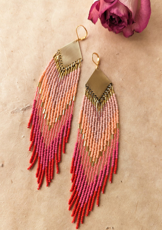 Moon & Milk handcrafted beaded earrings in ombré shades of rose, coral, and red with brass findings, displayed beside a dried pink rose on linen.