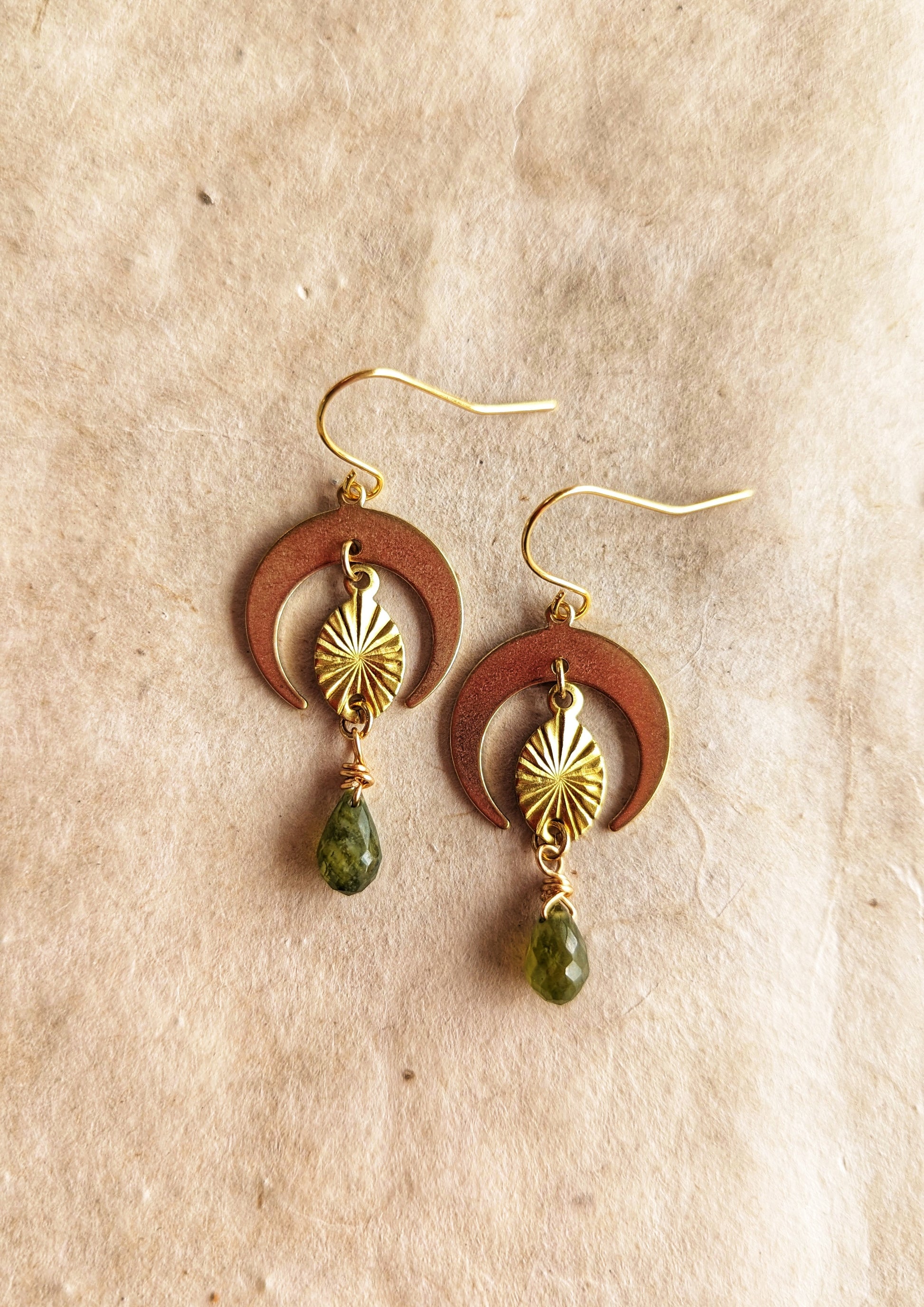 Moon & Milk -Gold brass earrings with crescent moon shapes, sunburst design, and dangling green garnet teardrop gemstones.  Beige, rustic background.