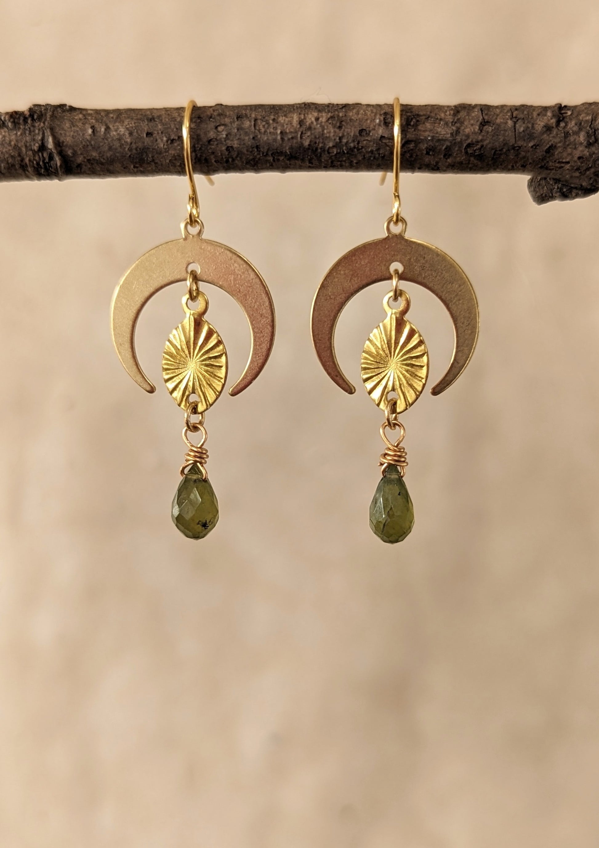 Moon & Milk -Gold brass earrings with crescent moon shapes, sunburst design, and dangling green garnet teardrop gemstones. Hanging on a branch, beige background.